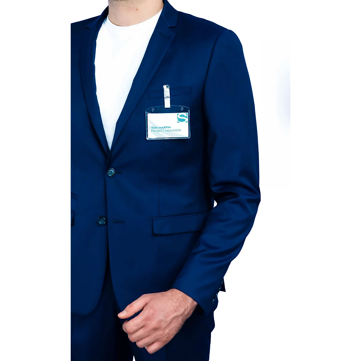 Un homme en costume bleu foncé avec un t-shirt blanc porte un badge nominatif sur sa poche poitrine.