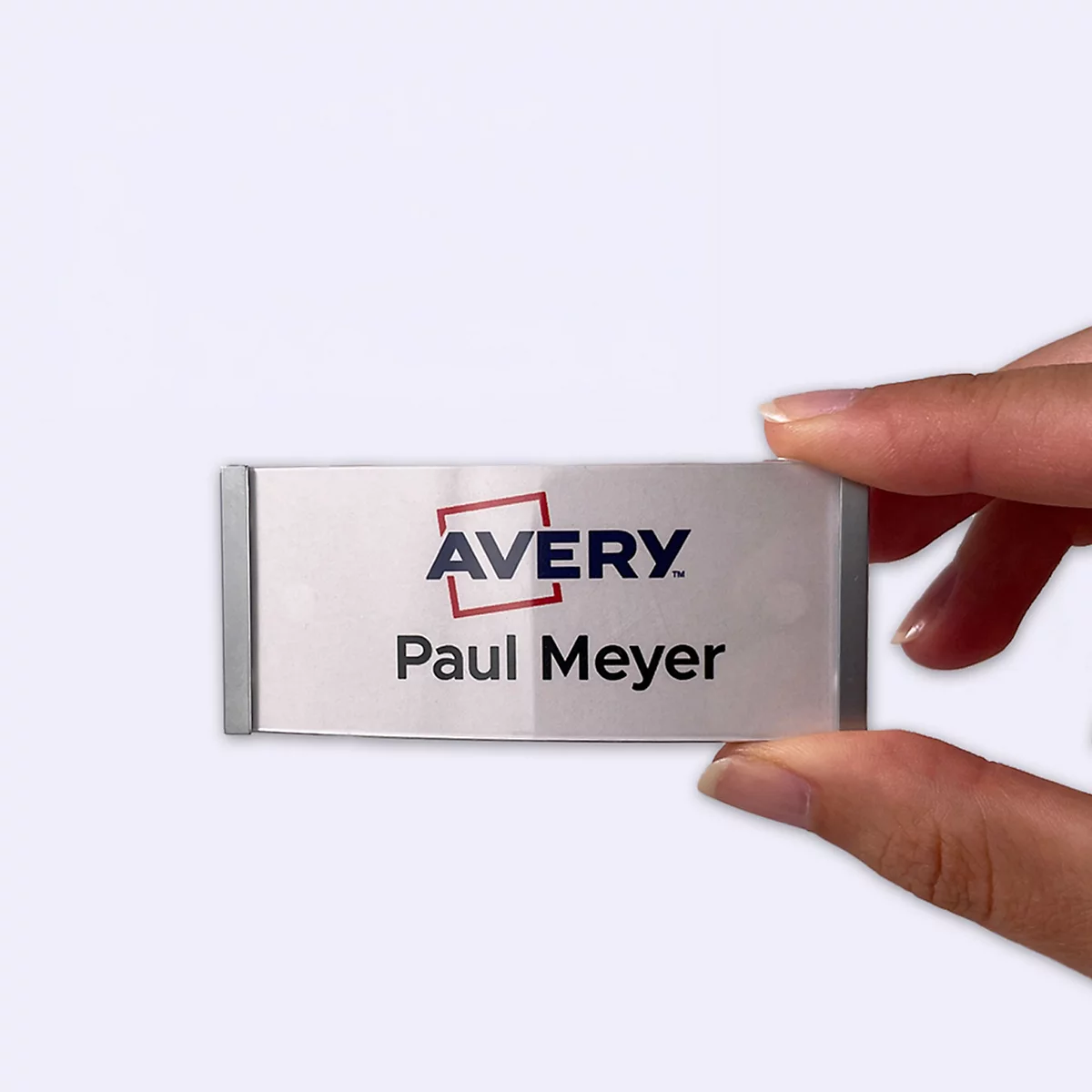 Een hand houdt een zilveren naamplaatje met het Avery-logo en de naam Paul Meyer vast tegen een witte achtergrond.