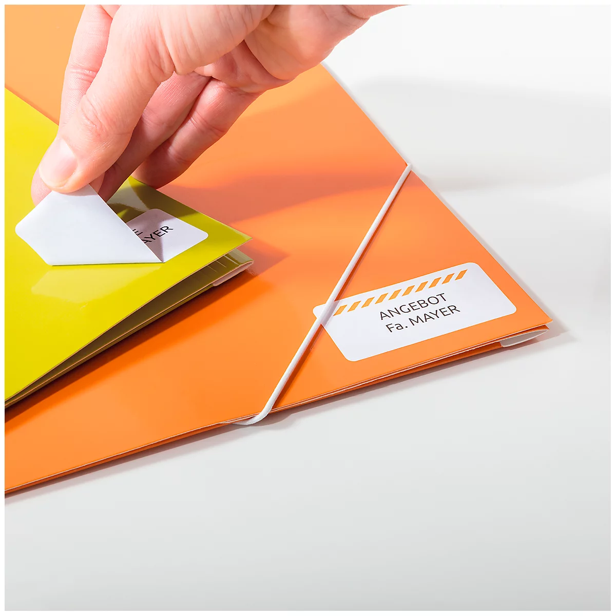 A hand peeling a sticker from a yellow folder while an orange folder lies next to it. Both folders have stickers with text.