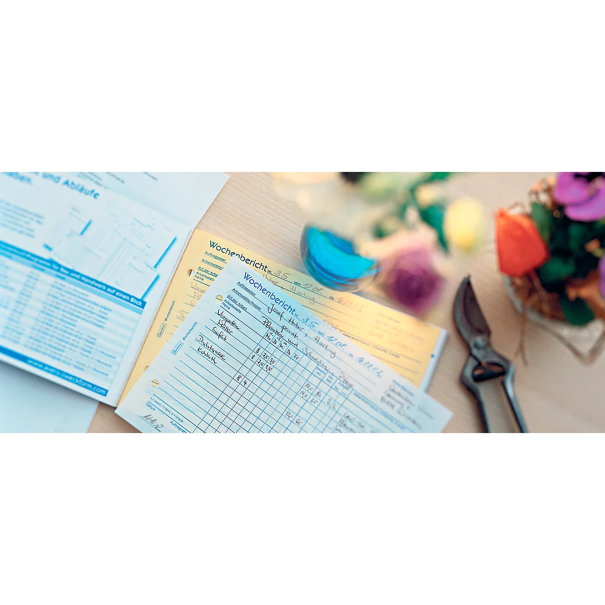 Close-up of notes, scissors and flowers on a wooden surface. The notes say 'Wochenbericht'.