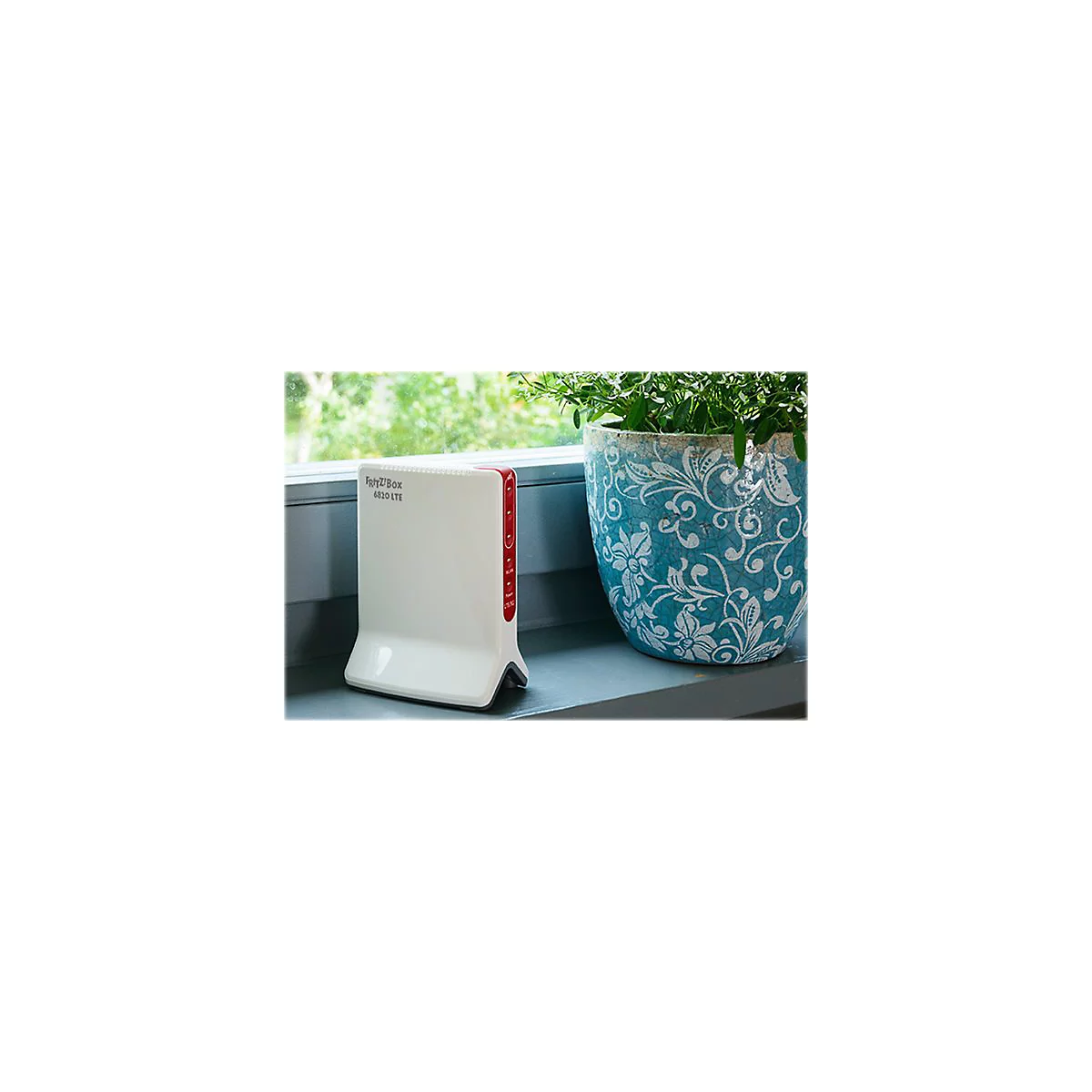 Fritz!Box 6820 LTE blanche sur un rebord de fenêtre. À droite, un pot avec des plantes vertes, derrière, un paysage vert flou.