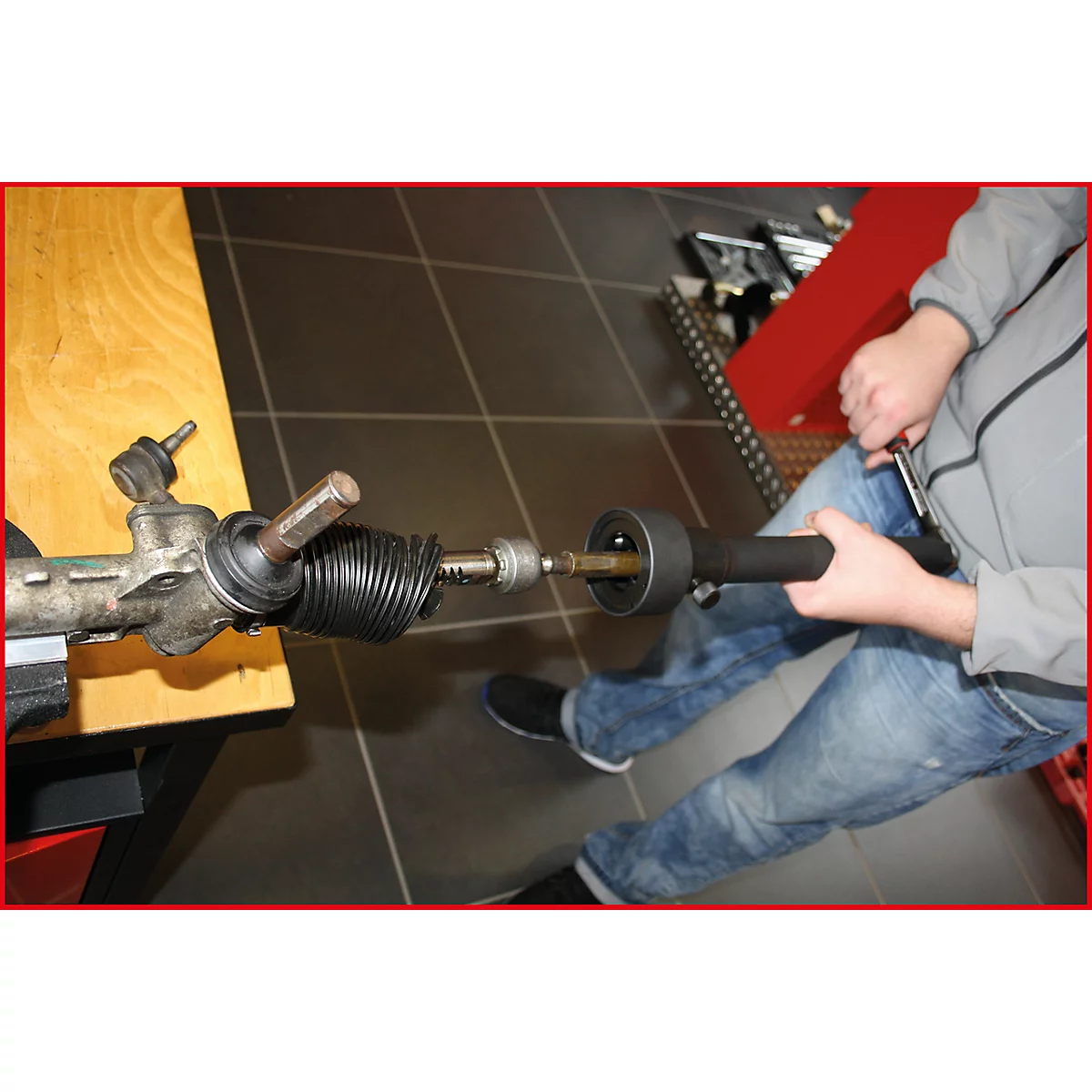 Mechaniker repariert das Lenkgetriebe eines Autos. Der Mann in Jeans und grauer Jacke schraubt an dem Bauteil.