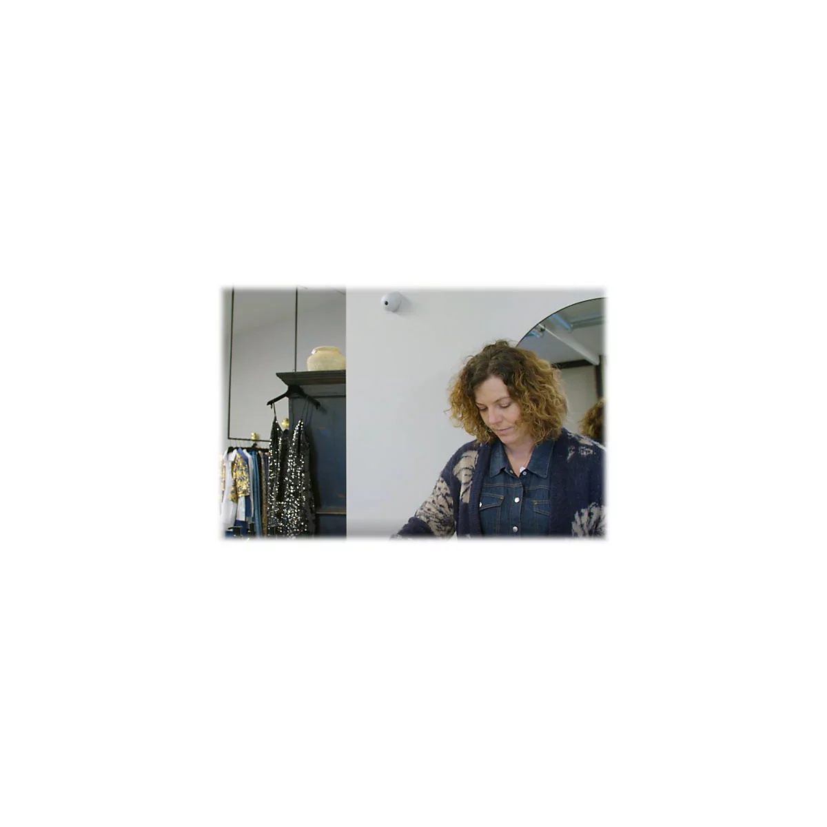 Eine Frau mit lockigem Haar betrachtet eine Kleidungsauswahl in einem Geschäft. Im Hintergrund Kleider, Spiegel.