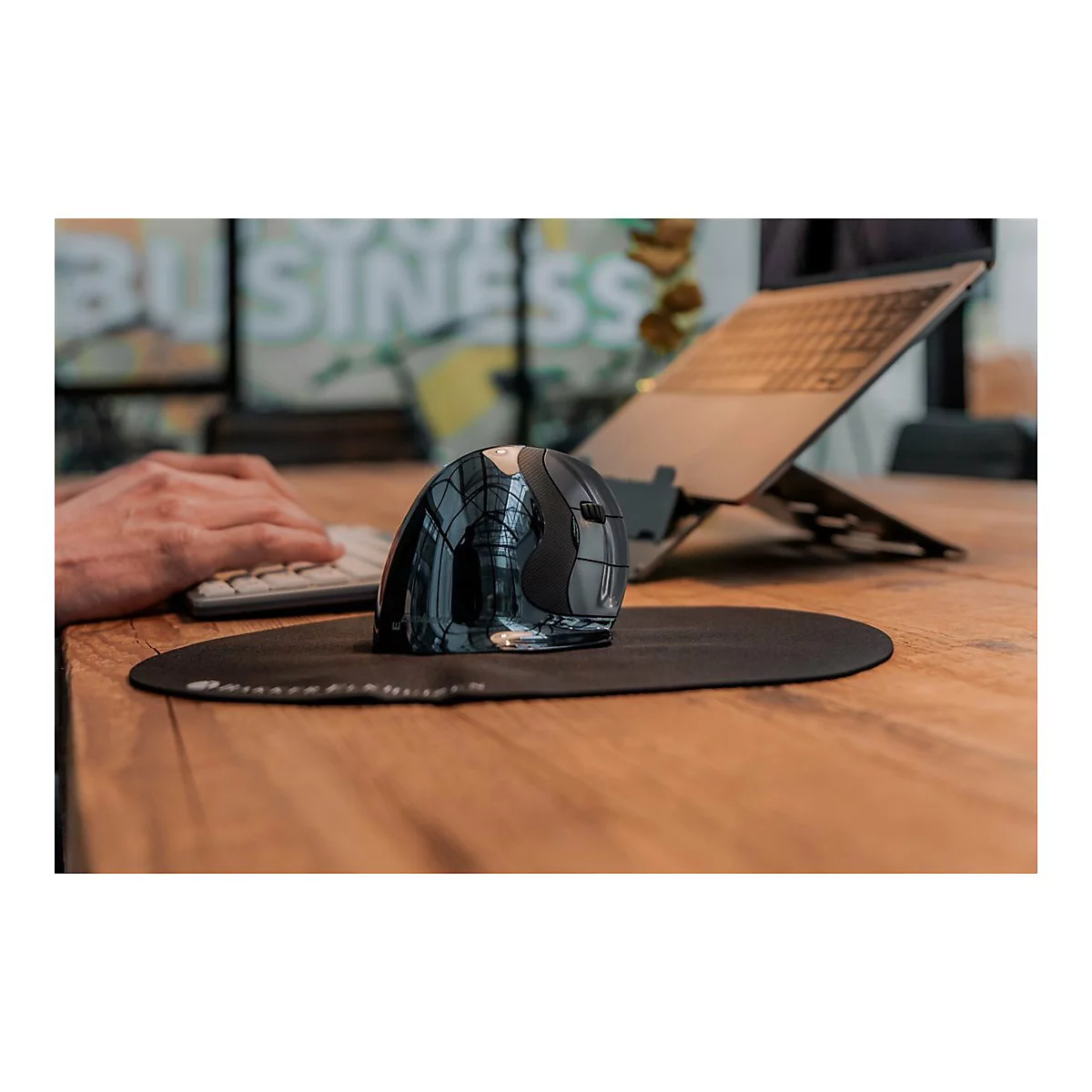 Ein ergonomische Maus liegt auf einem schwarzen Mauspad, daneben ein Laptop auf einem Ständer. Eine Hand bedient eine Tastatur.