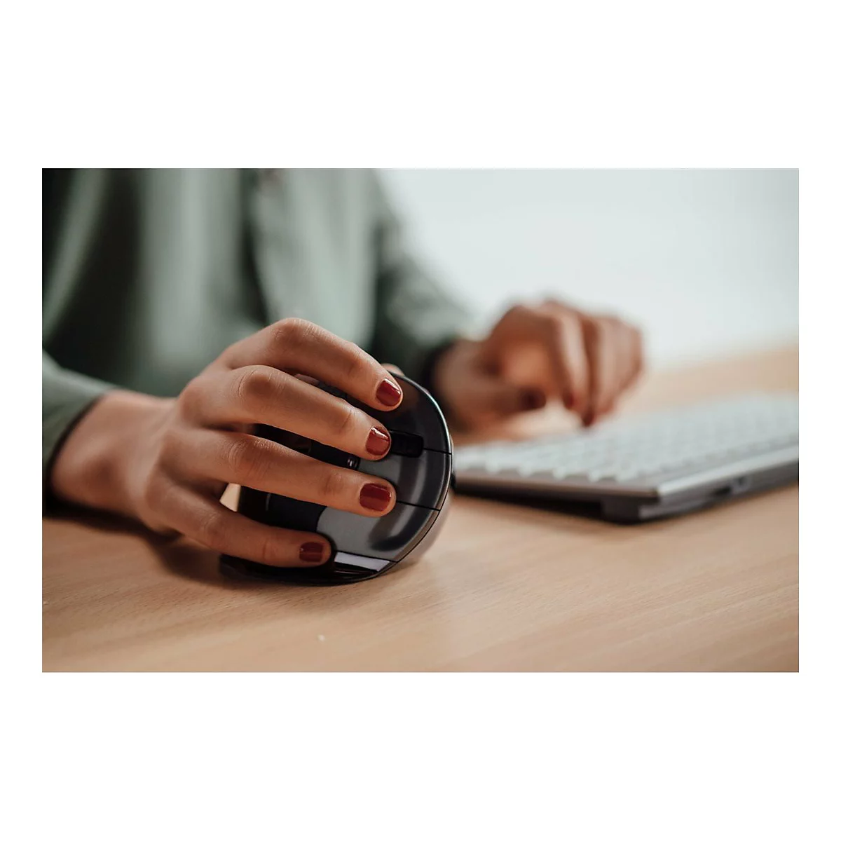 Person, die eine ergonomische Maus benutzt; rote Fingernägel auf Holzoberfläche. Eine Tastatur ist im Hintergrund.