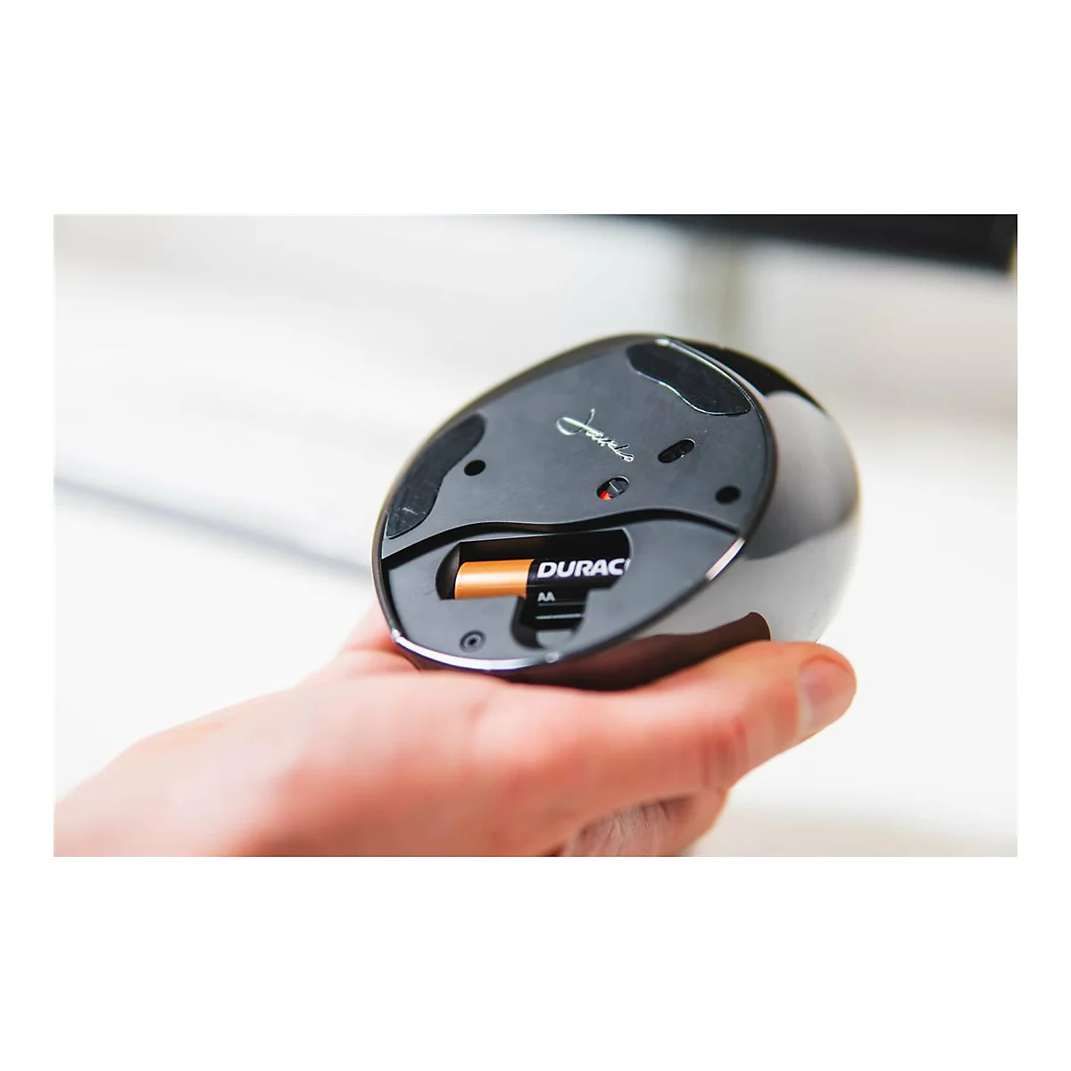 Ein Mann hält eine schwarze Computermaus. Man sieht die Unterseite mit einer eingelegten Batterie. Auf der Batterie steht Duracell.
