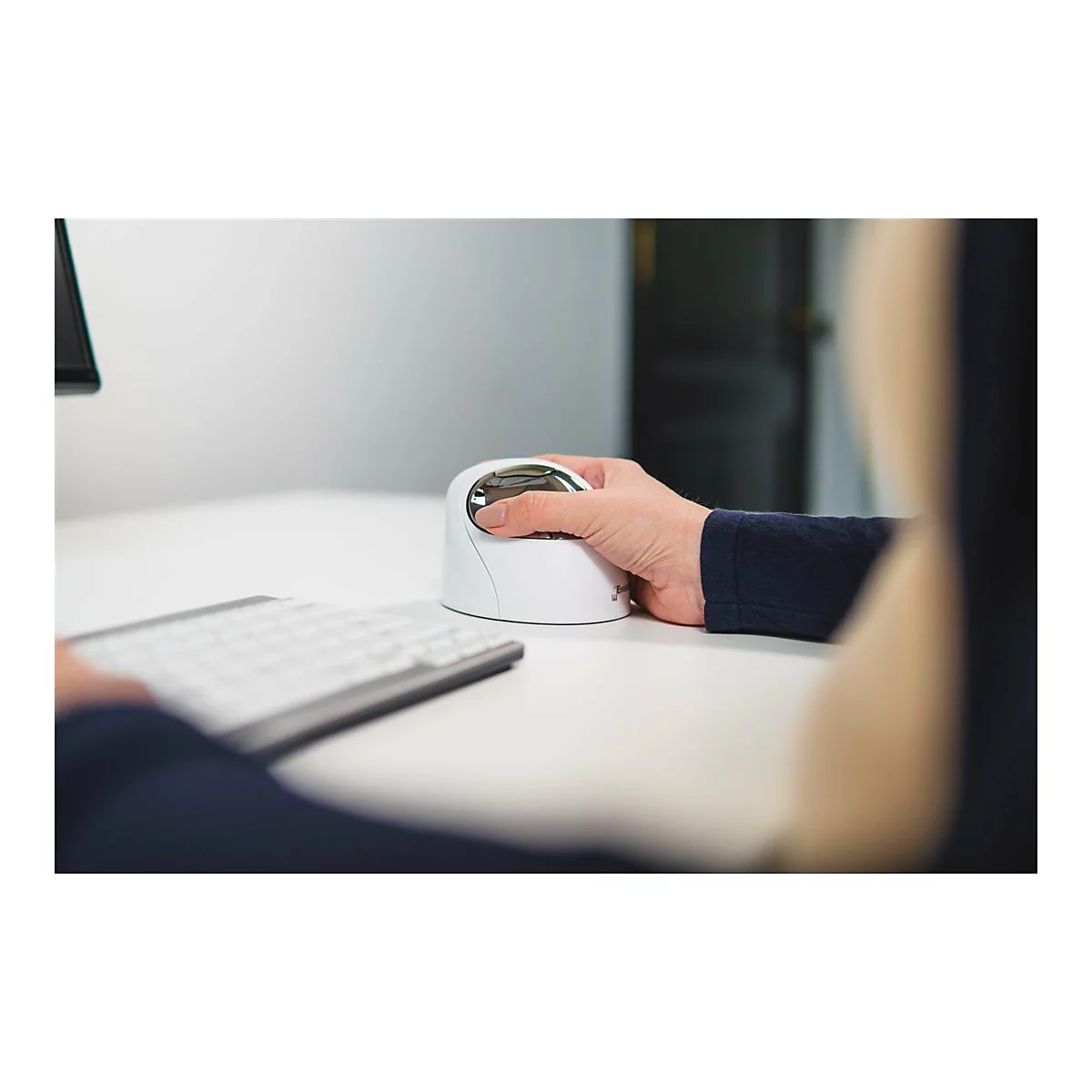 Person bedient vertikale Maus. Weiße Maus, silbernes Oberteil, auf weißem Tisch. Dunkler Pullover, Tastatur, heller Hintergrund.
