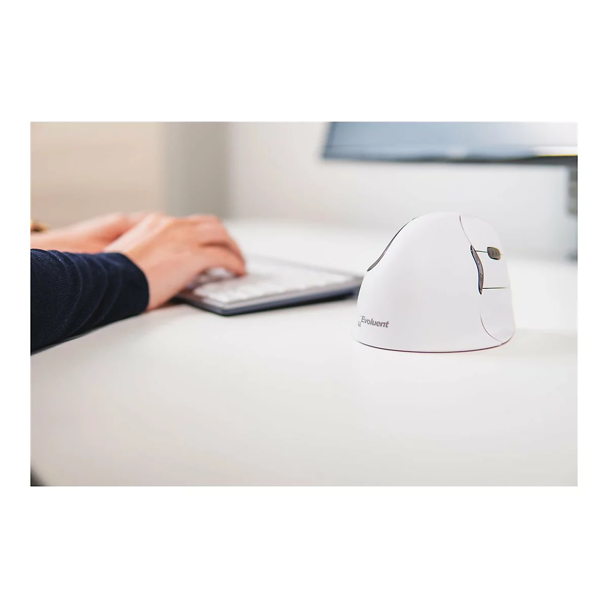 Weisse Vertikalmaus, neben Person die auf Tastatur tippt, auf weissem Tisch. Im Hintergrund Monitor.