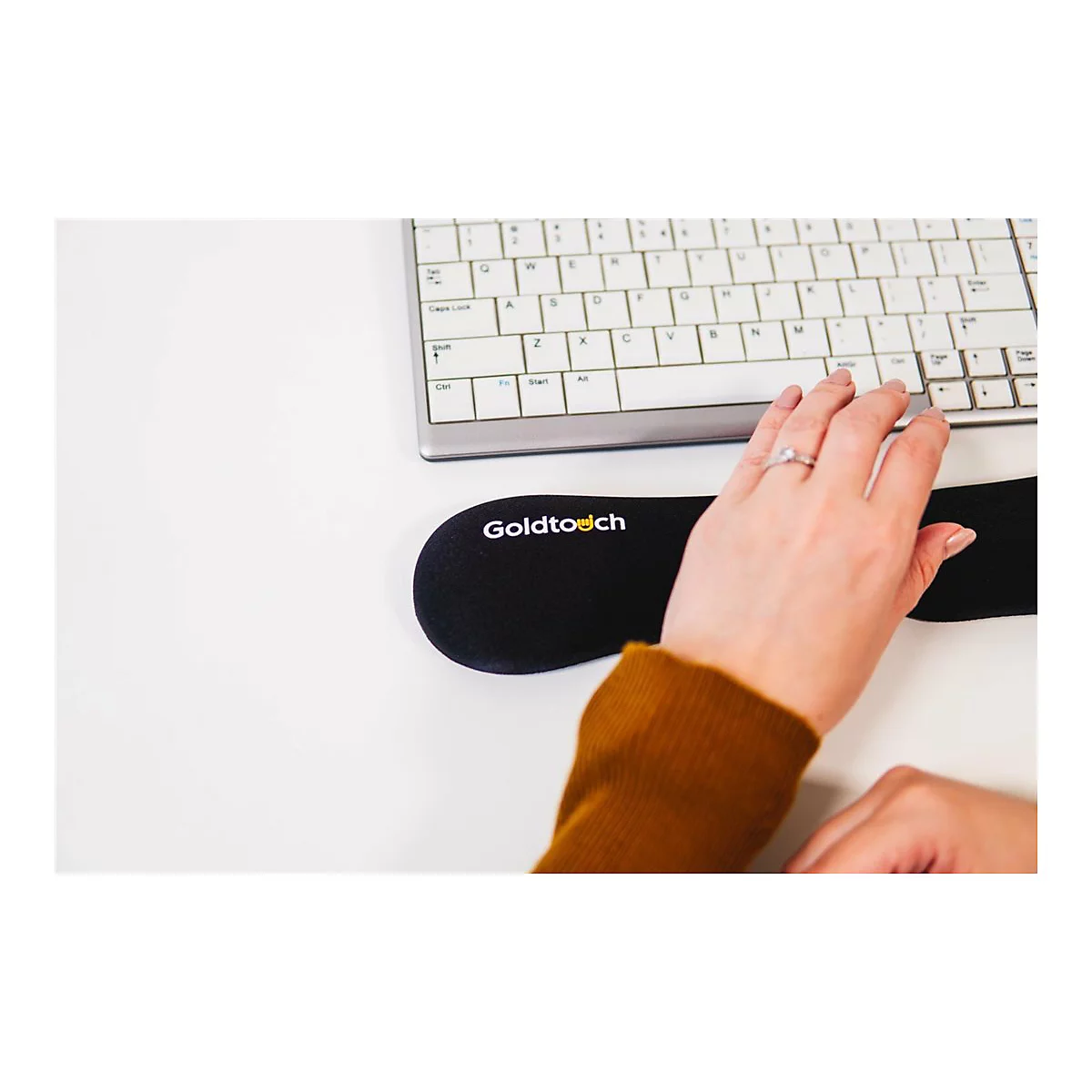 Eine Hand mit Ring auf einer schwarzen Handgelenkauflage mit Goldtouch-Logo, neben einer Tastatur. Brauner Pullover im unteren Bereich.
