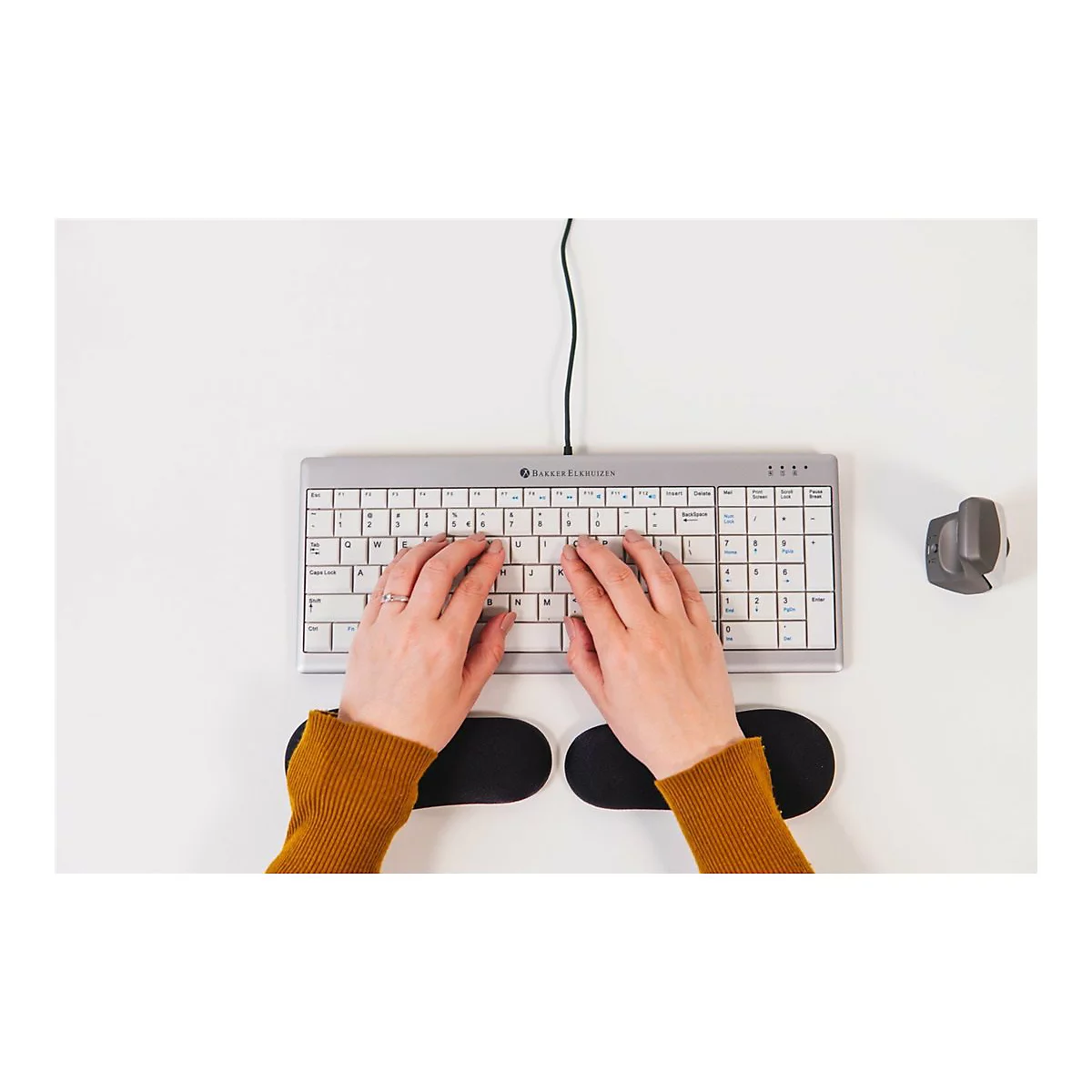 Person tippt auf einer ergonomischen Tastatur. Auf dem weißen Schreibtisch liegen eine Maus und Handgelenkstützen.