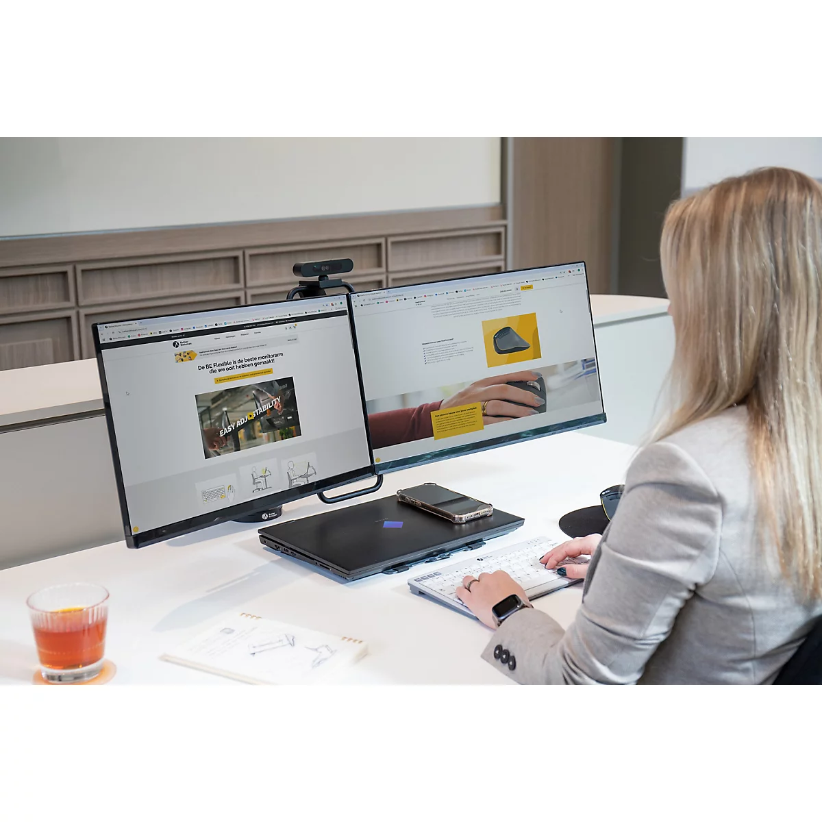 Eine Frau arbeitet an einem Schreibtisch mit zwei Bildschirmen. Sie tippt auf einer Tastatur, ein Cocktail steht neben ihr.