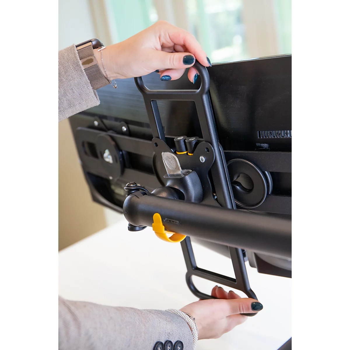 Een persoon houdt een zwarte monitorbeugel vast, gele band, donkerblauwe nagellak.