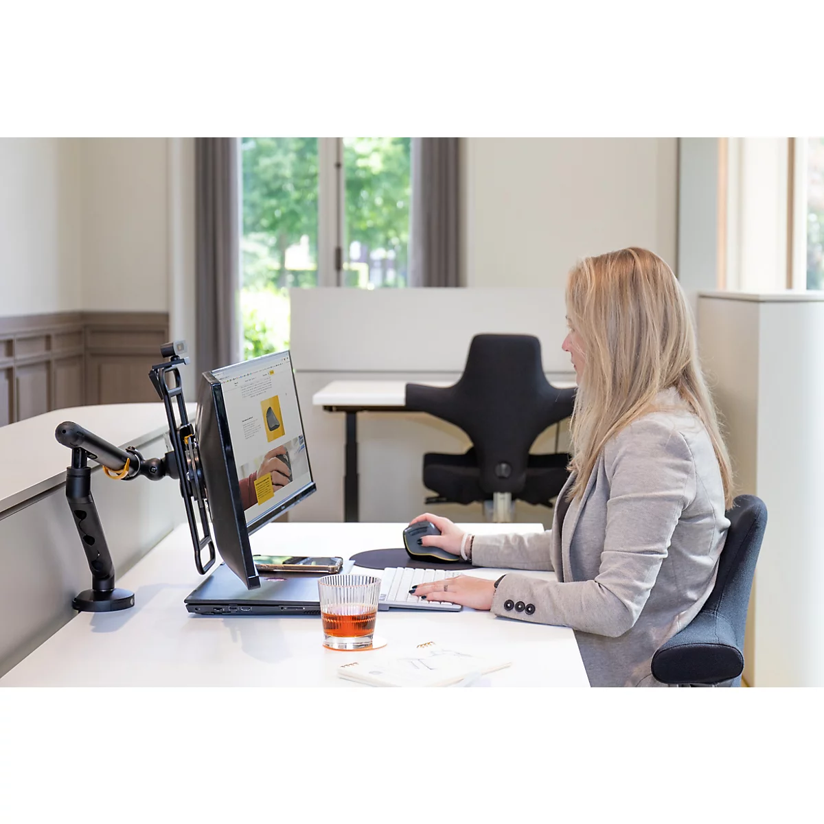 Een vrouw werkt aan een bureau. Ze gebruikt een computer, een in hoogte verstelbare monitorarm en een ergonomische muis.