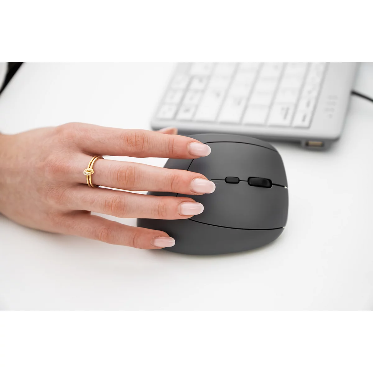 Hand mit Ring, die eine graue ergonomische Maus bedient, Tastatur im Hintergrund.