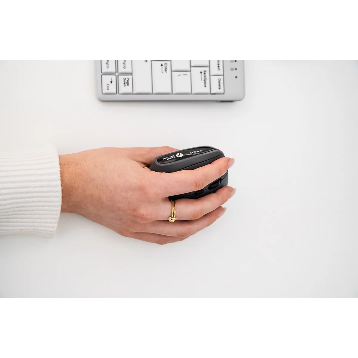Eine Person hält eine schwarze Maus. Auf dem Tisch liegt eine Tastatur und ein goldener Ring am Finger.