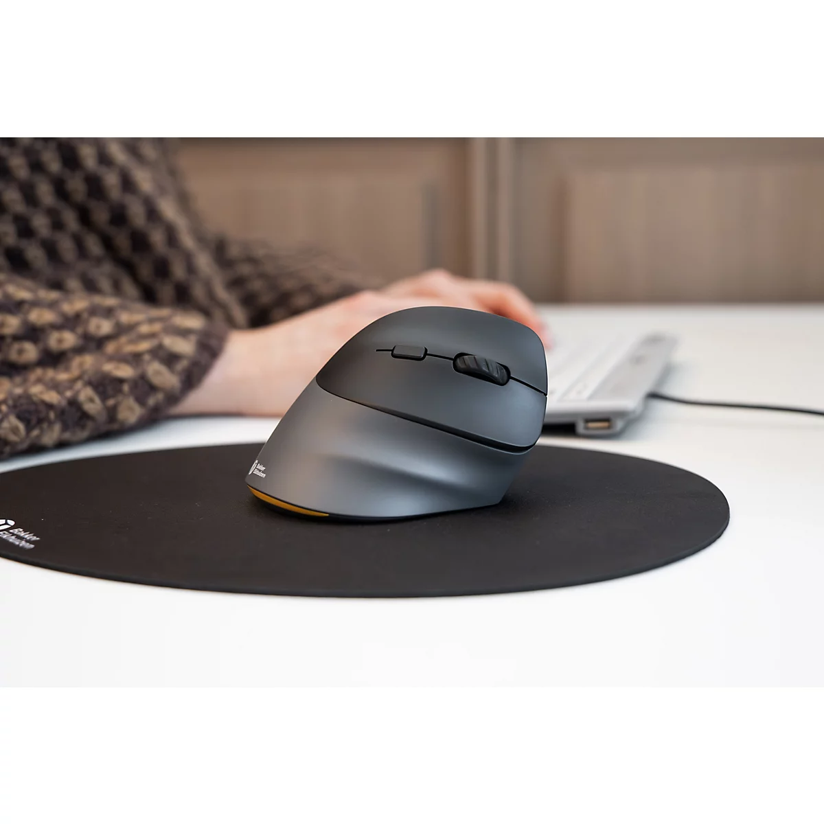 Eine ergonomische, graue Vertikalmaus steht auf einem runden, schwarzen Mauspad, während im Hintergrund jemand tippt.