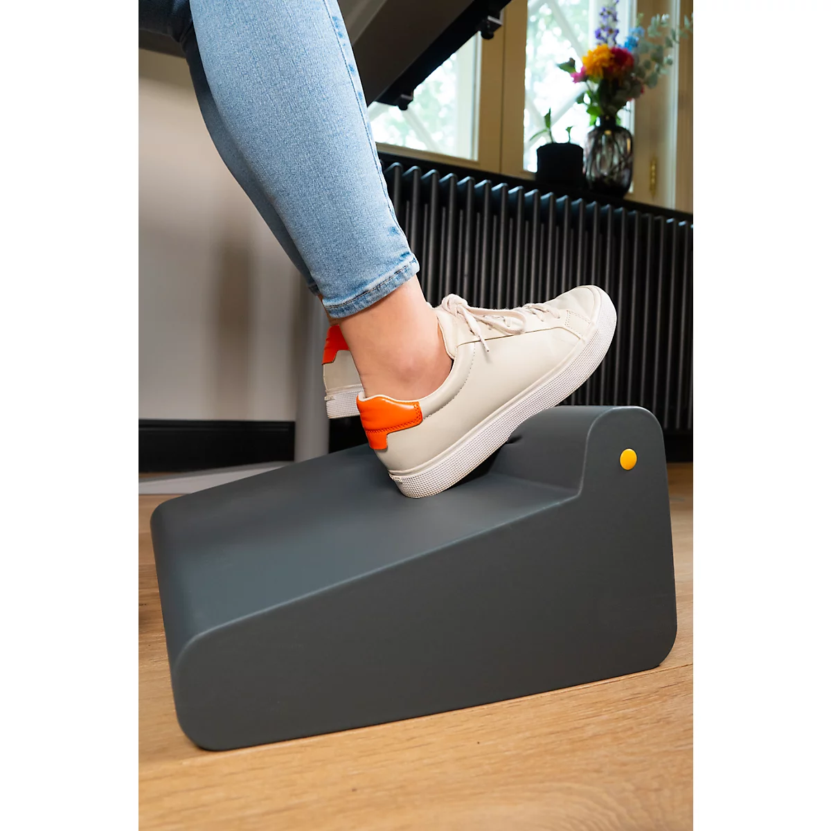Une personne pose un pied sur un repose-pieds gris. La chaussure est blanche avec un détail orange.