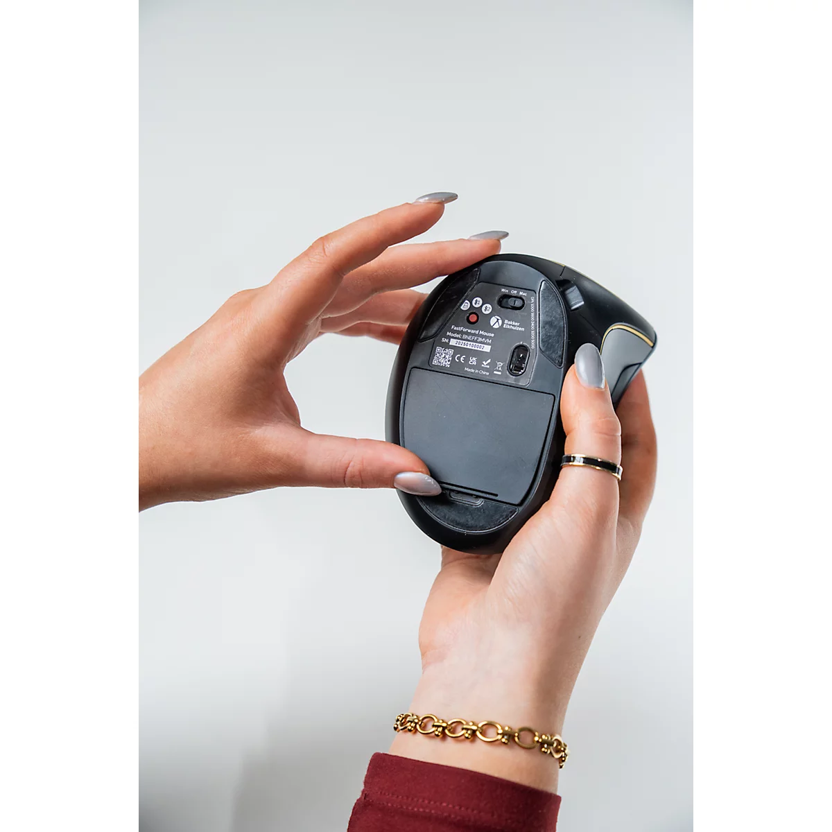 Hände halten eine schwarze, ergonomische Maus, Details der Unterseite. Ringe und Armband sind sichtbar.