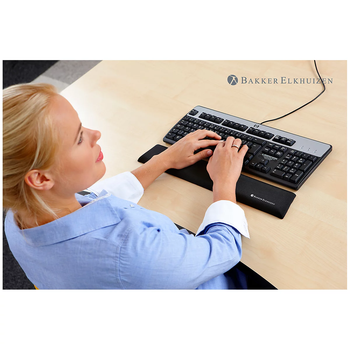 A woman typing on a keyboard. She has blonde hair, is wearing a blue shirt, and is sitting at a light-colored desk.
