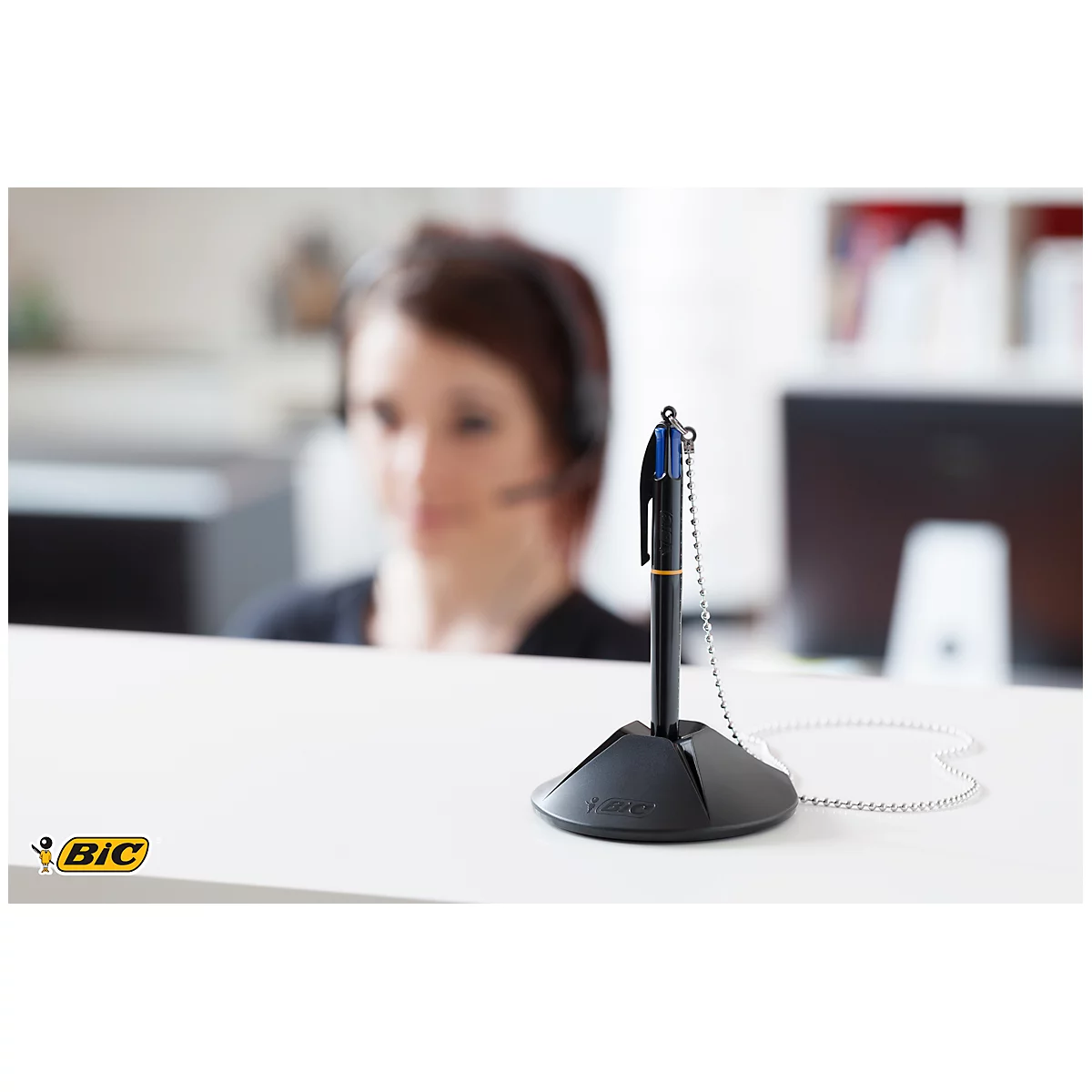 Pen with chain in a pen holder. In the background a woman with a headset at the desk.