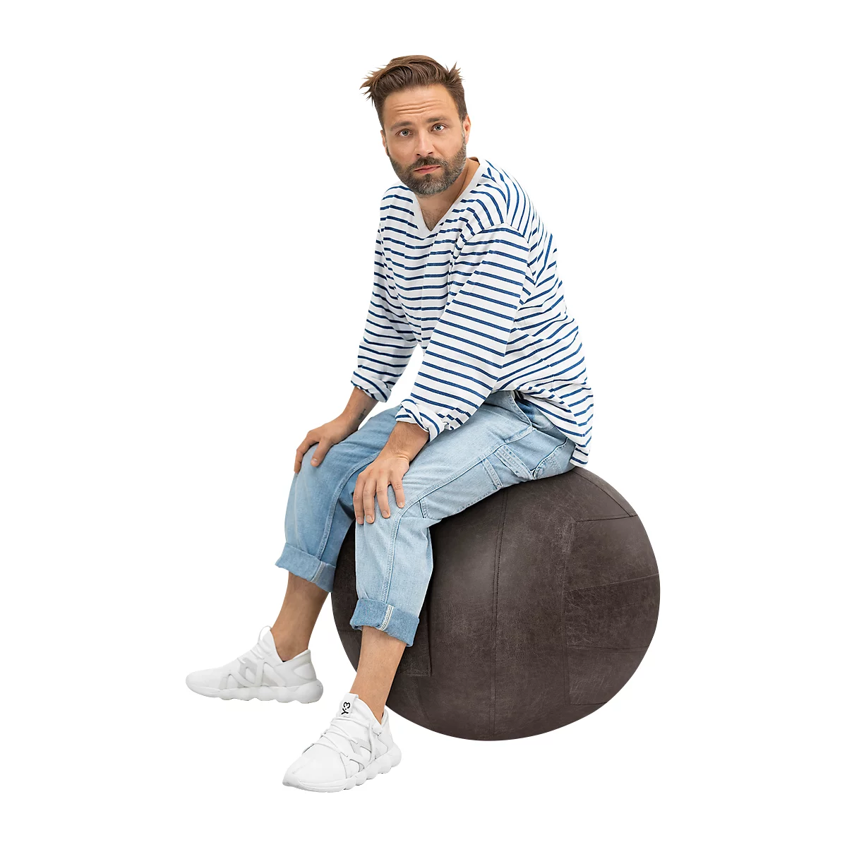 Un hombre sentado en un balón de ejercicio redondo y marrón. Lleva vaqueros, una camisa a rayas y zapatillas blancas.