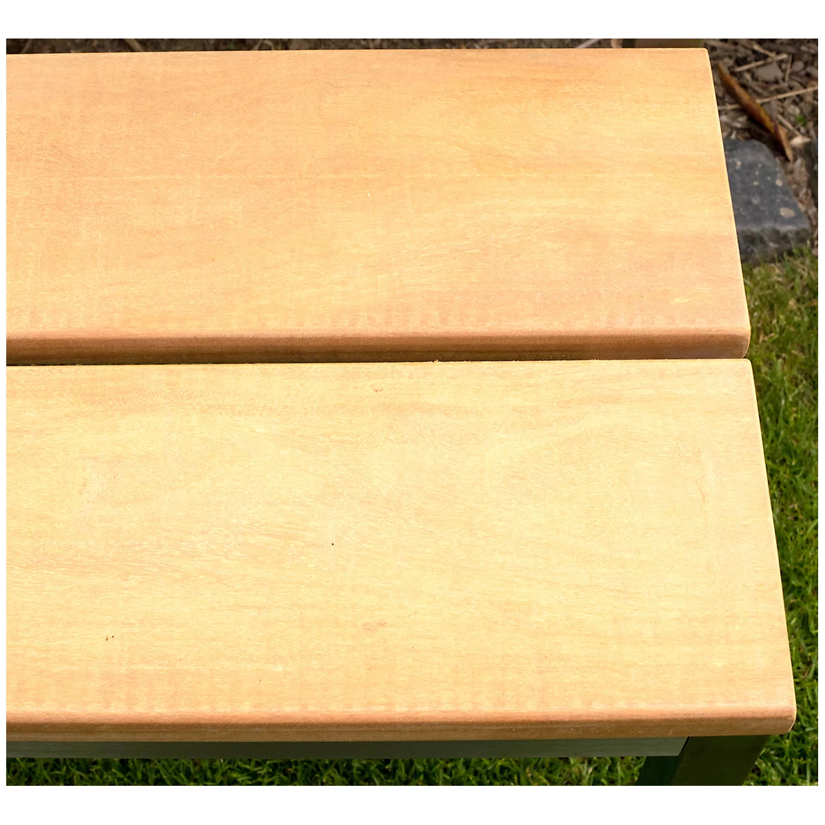 Gros plan sur un banc en bois brun clair avec des bords arrondis. En arrière-plan, on voit de l'herbe et un mur de pierre grise.