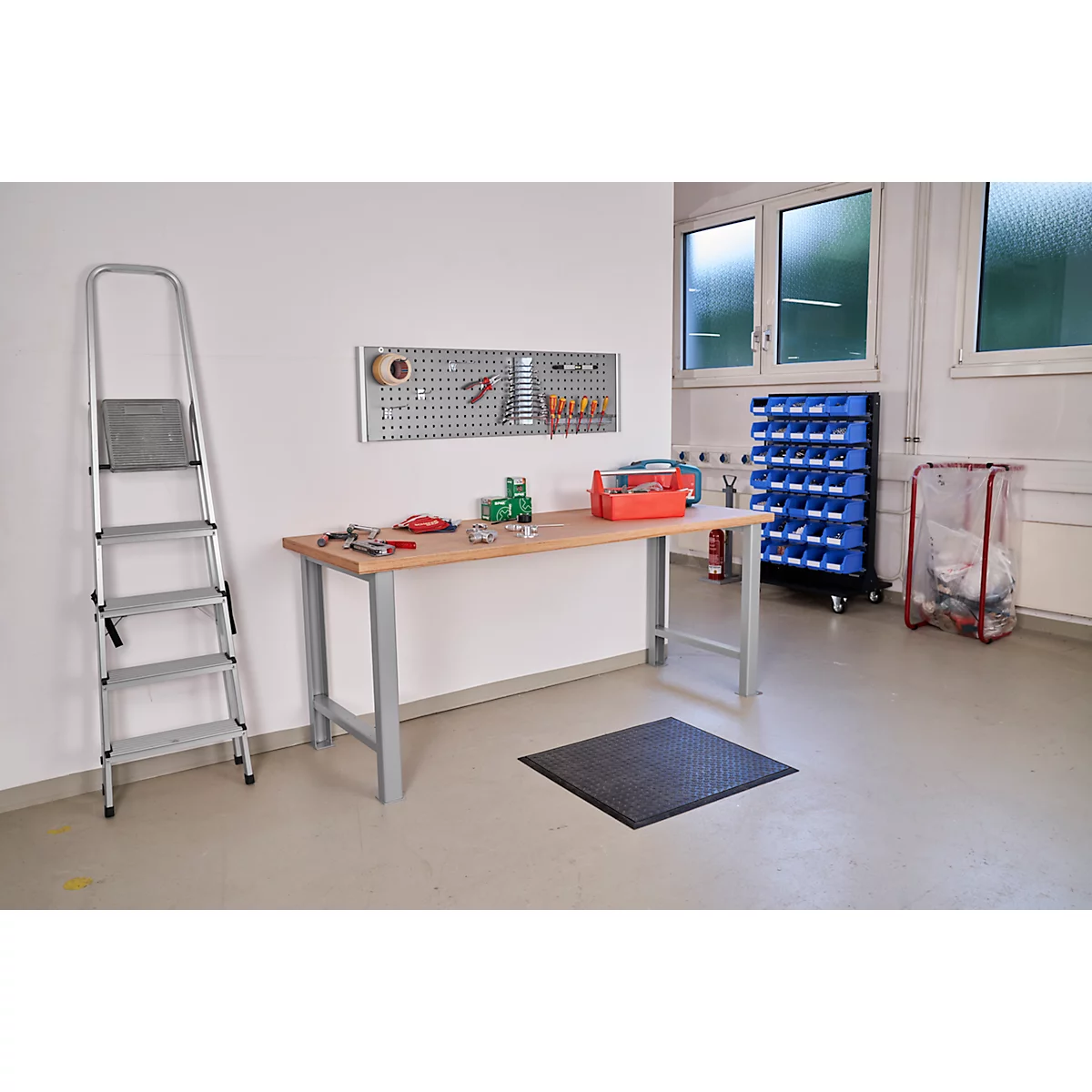Taller: banco de trabajo, panel perforado con herramientas, escalera, cajas azules, caja de herramientas, alfombra de trabajo. Ventanas en el fondo.