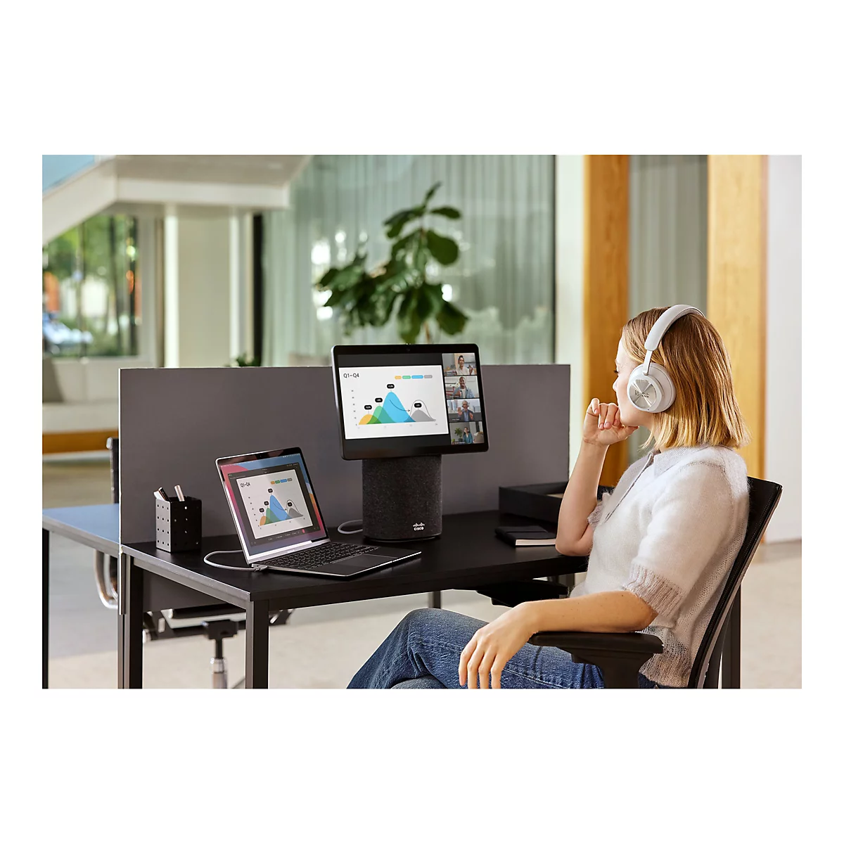 Eine Frau sitzt an einem Schreibtisch mit Headset und schaut auf ein Tablet mit Videokonferenz. Auf dem Tisch stehen ein Laptop und ein zylinderförmiger Lautsprecher.