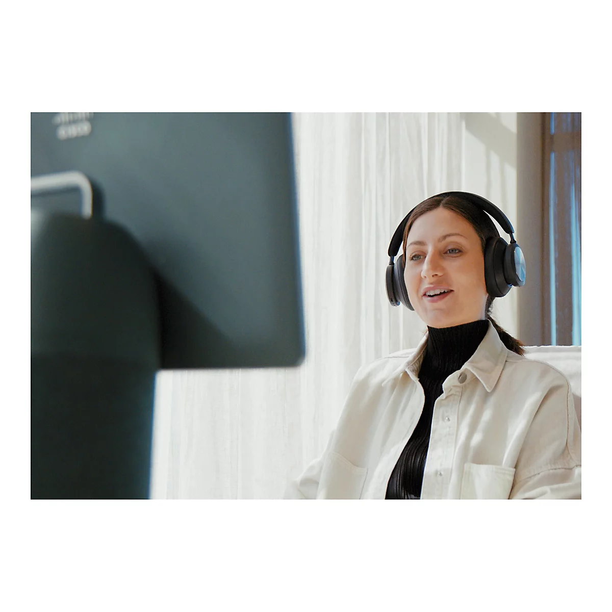 Eine Frau mit Kopfhörern blickt lächelnd auf einen Bildschirm. Sie trägt ein schwarzes Oberteil und eine helle Jacke.