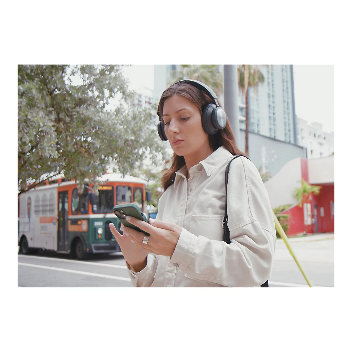 Eine Frau mit Kopfhörern benutzt ihr Handy. Im Hintergrund ein Bus, Büsche und Gebäude.