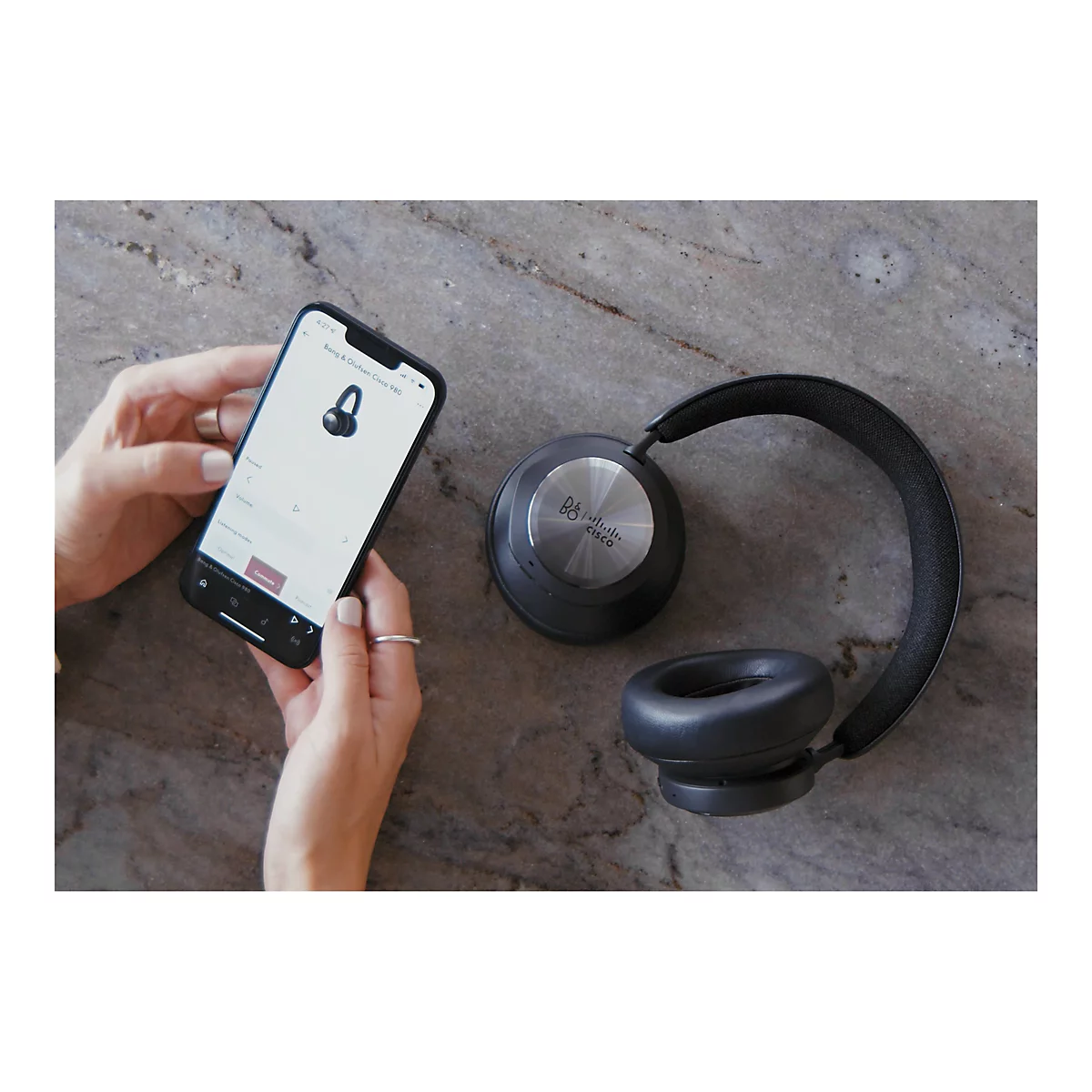 Nahaufnahme von Kopfhörern und einem Smartphone auf marmoriertem Untergrund. Eine Hand hält das Telefon, das die Einstellungen der Kopfhörer anzeigt. Die Kopfhörer sind schwarz und haben einen silberfarbenen Mittelteil. Auf dem Telefon steht "Bang & Ol...