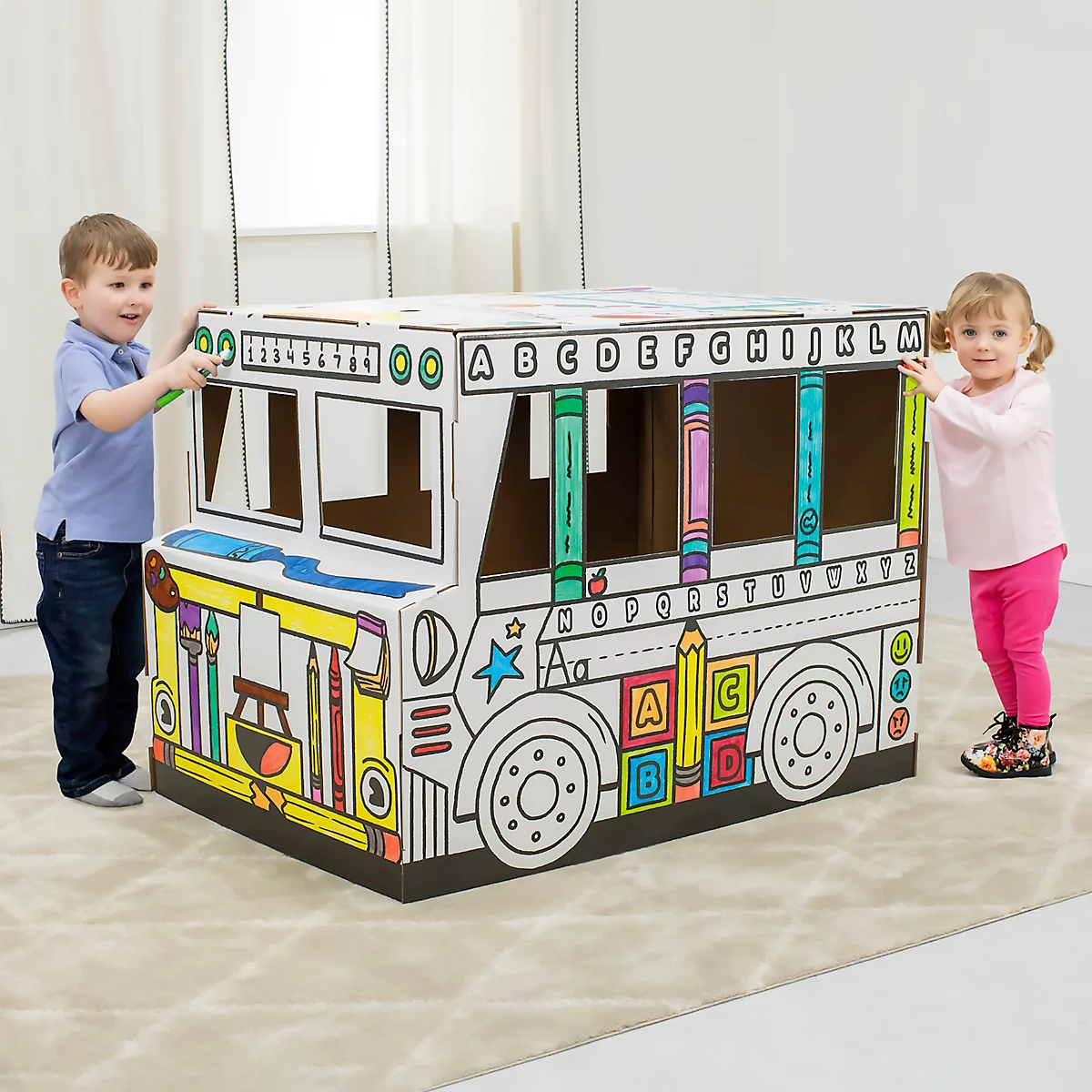 Deux enfants se tiennent debout près d'un grand bus en carton peint. Le bus est décoré de lettres et de crayons de couleur. Scène joyeuse !