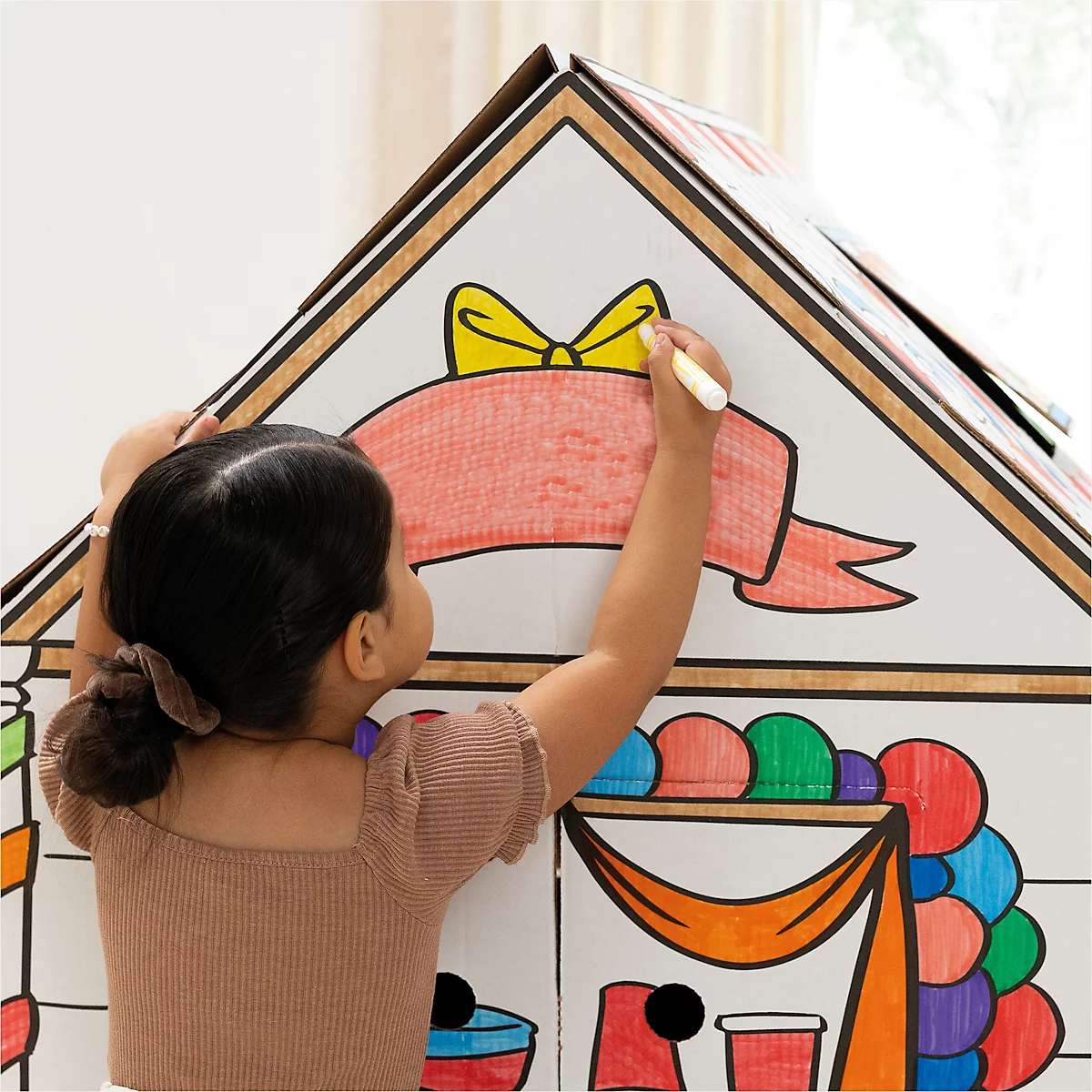 Un enfant colorie une maison en carton avec un stylo. Il y a un nœud et une bannière rouge sur la maison.