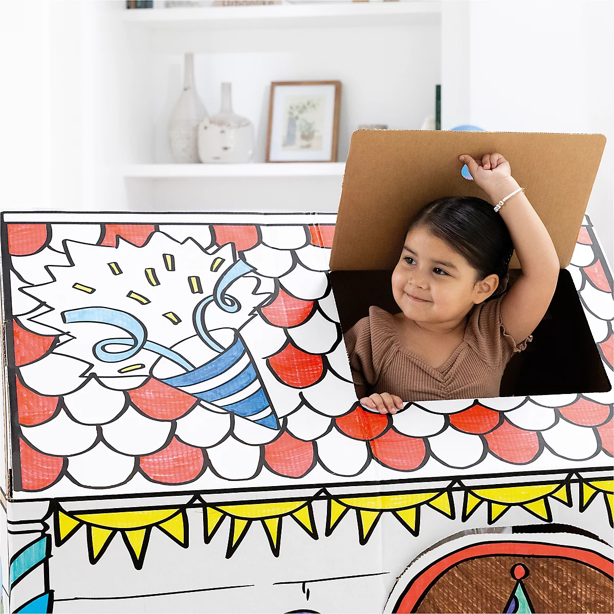 Une petite fille souriante regarde à l'extérieur d'une maison en carton faite maison. La maison est décorée de dessins colorés. Cela semble joyeux.