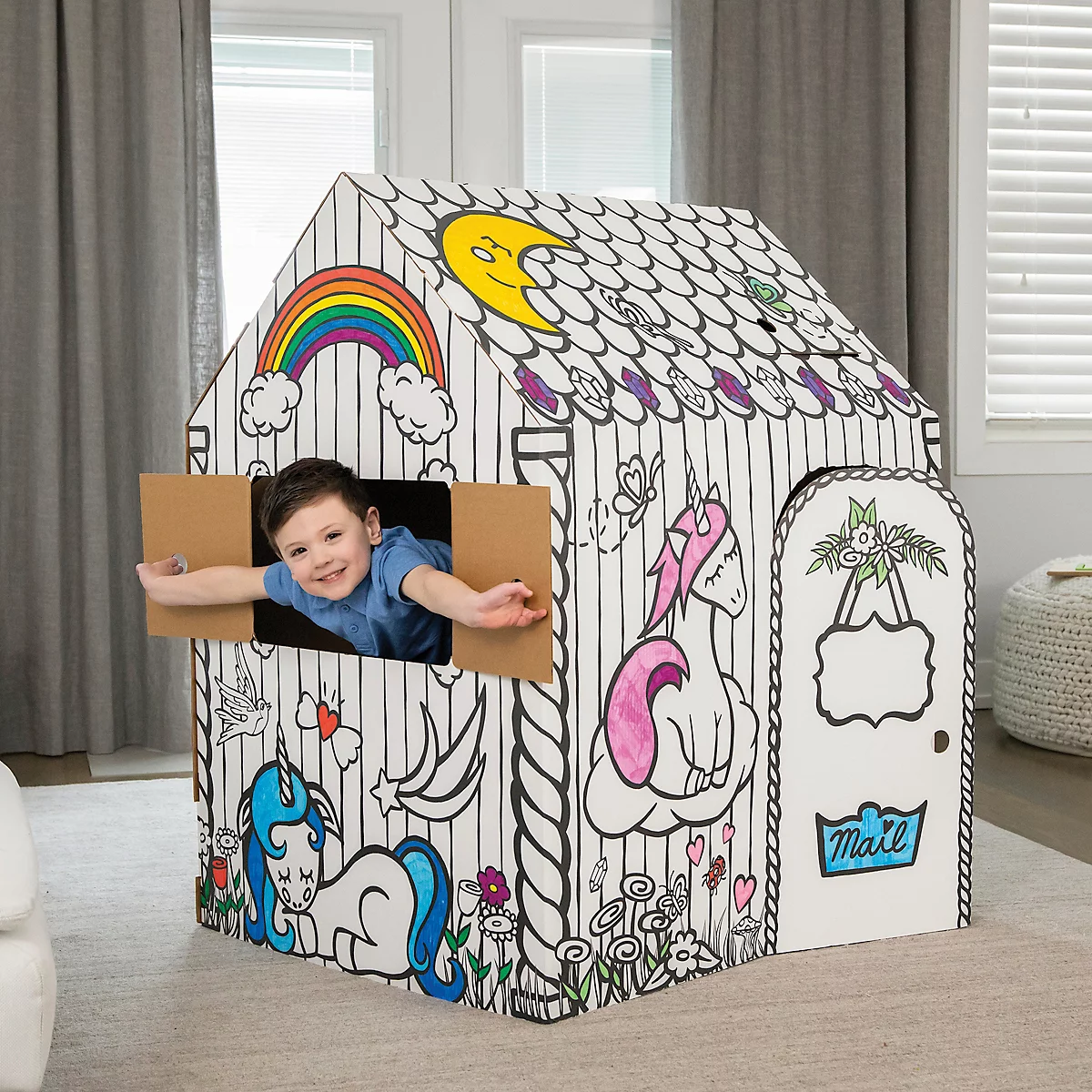 Un garçon se penche par la fenêtre d'une maison de jeu peinte avec des licornes et des arcs-en-ciel. La maison est en carton.