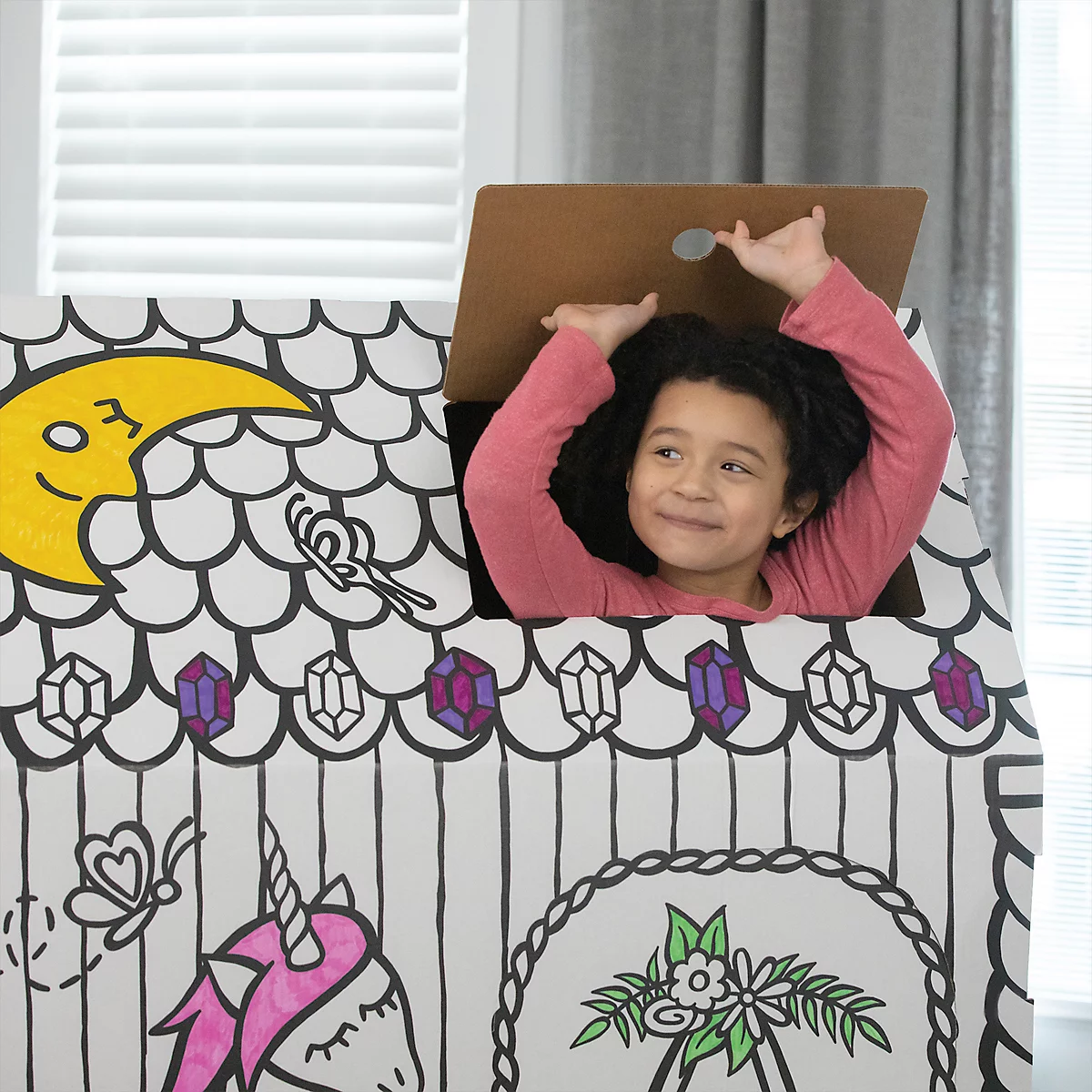 Un enfant souriant regarde à l'extérieur d'une maison en carton décorée de dessins de licornes et de lunes. L'enfant lève les bras.