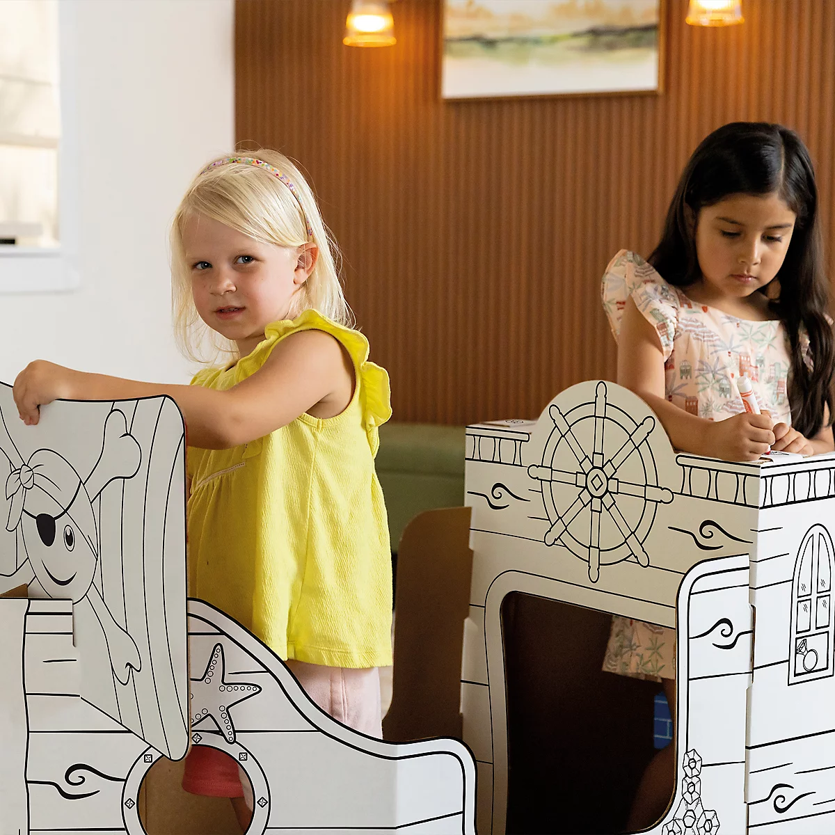 Zwei Mädchen spielen in einem Pappschiff, eines mit Augenklappe und Stern, das andere am Steuerrad.