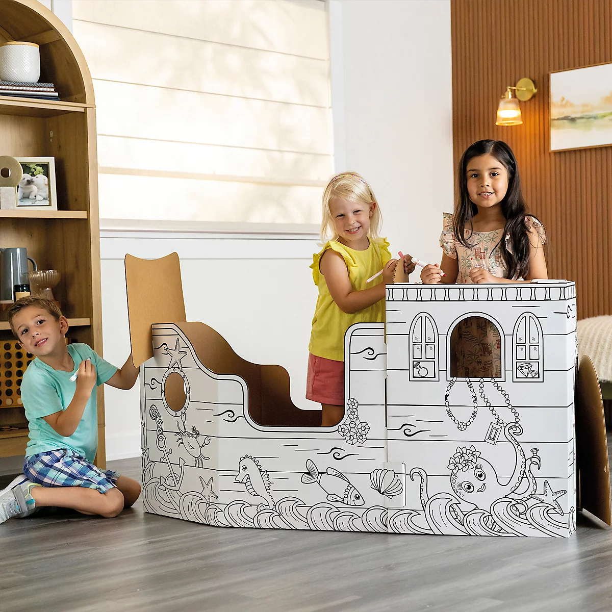 Drei Kinder spielen in einem bemalten Pappschiff. Das Schiff ist von einem Kind bemalt, mit Tintenfisch, Fischen und Wellen.