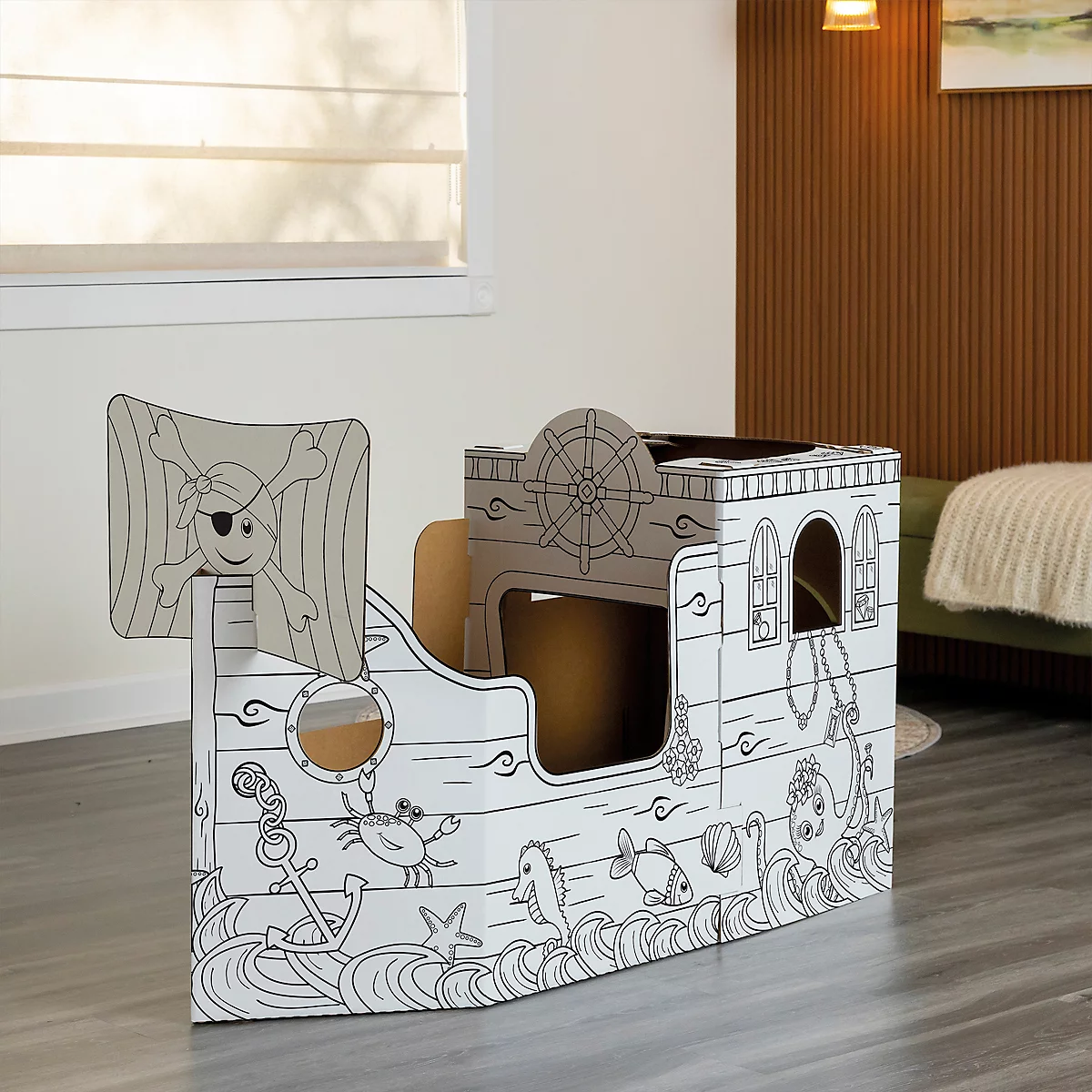 Une cabane de jeu en forme de bateau pirate en carton avec une voile de pirate, une ancre, des poissons et des vagues en guise de décoration. Il se trouve dans une pièce.