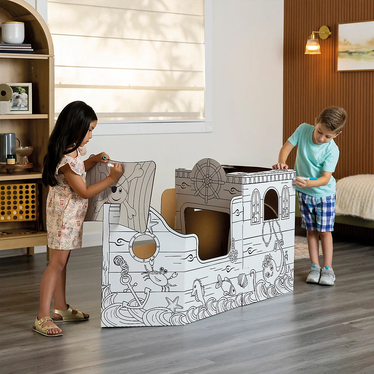 Deux enfants colorient un bateau pirate en carton. Une fille peint sur la voile, un garçon sur la poupe.