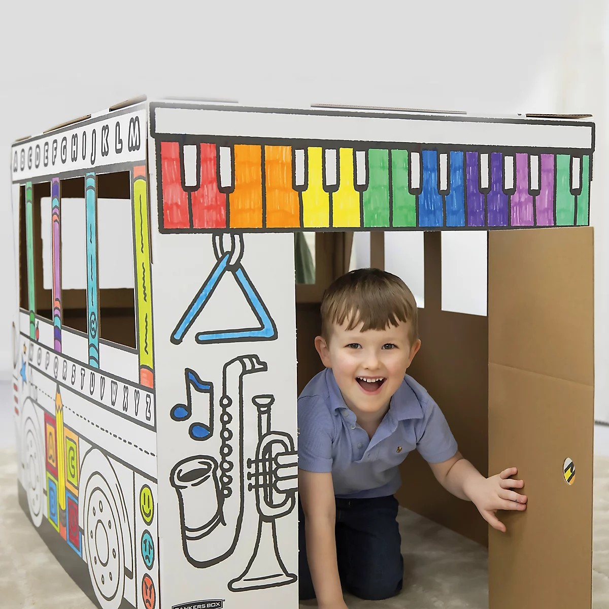 Ein lächelnder Junge in einem bemalten Spielbus aus Pappe. Auf dem Dach ist eine farbige Klaviatur abgebildet. Auf der Seite sind Musikinstrumente zu sehen.