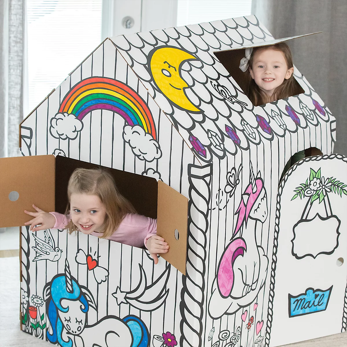 Zwei Mädchen spielen in einem bemalten Spielhaus. Das Haus hat ein Regenbogen-, Einhorn- und Mond-Design. Die Kinder lächeln.