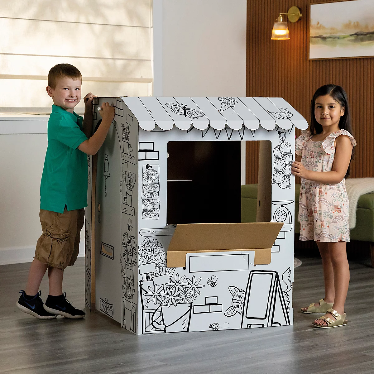 Deux enfants se tiennent à côté d'une maison en carton. La maison est décorée de dessins.