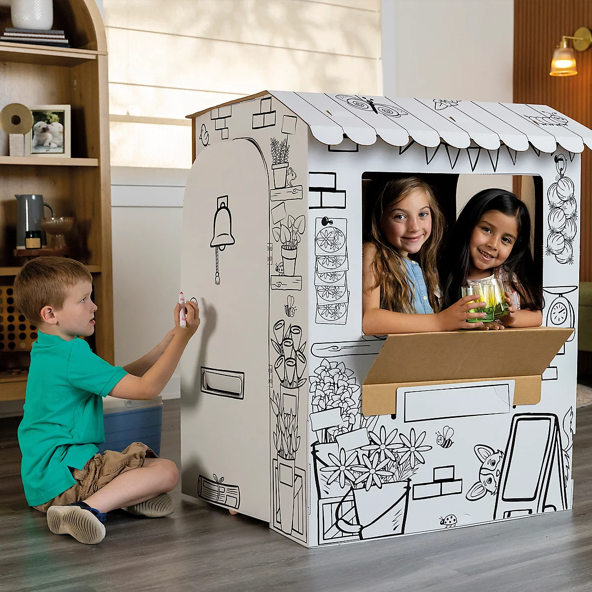 Un enfant dessine sur une boutique de jouets. Deux filles sont assises à la fenêtre et sourient. L'étal est dessiné sur du carton.