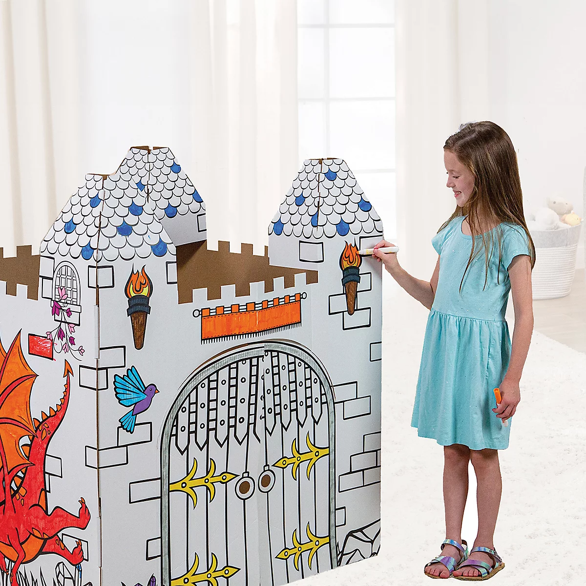 Ein Mädchen bemalt ein Pappschloss. Das Schloss ist mit Drachen, Türmen und einem Tor verziert. Es trägt ein blaues Kleid und einen Stift in der Hand.