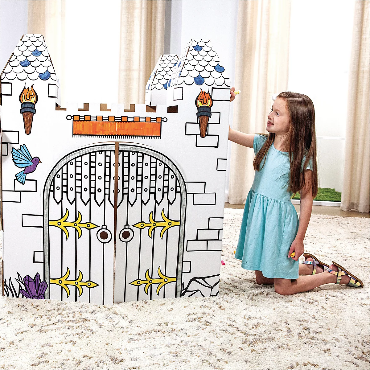 Fille agenouillée à côté d'un château en carton à peindre. Château avec créneaux, porte et tours. Jeune femme porte une robe bleu clair.