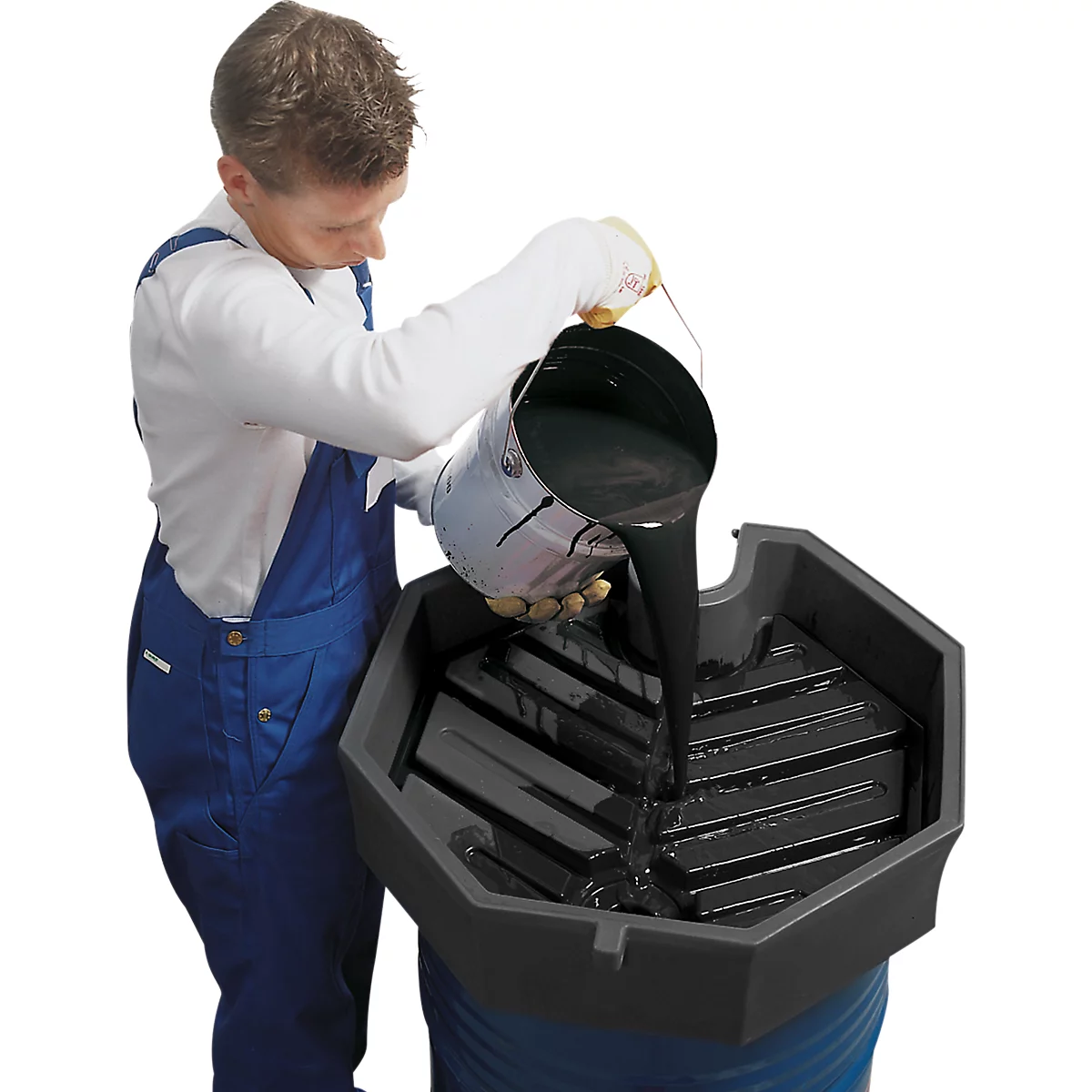 A man in blue overalls pours black liquid from a bucket into a drip tray.