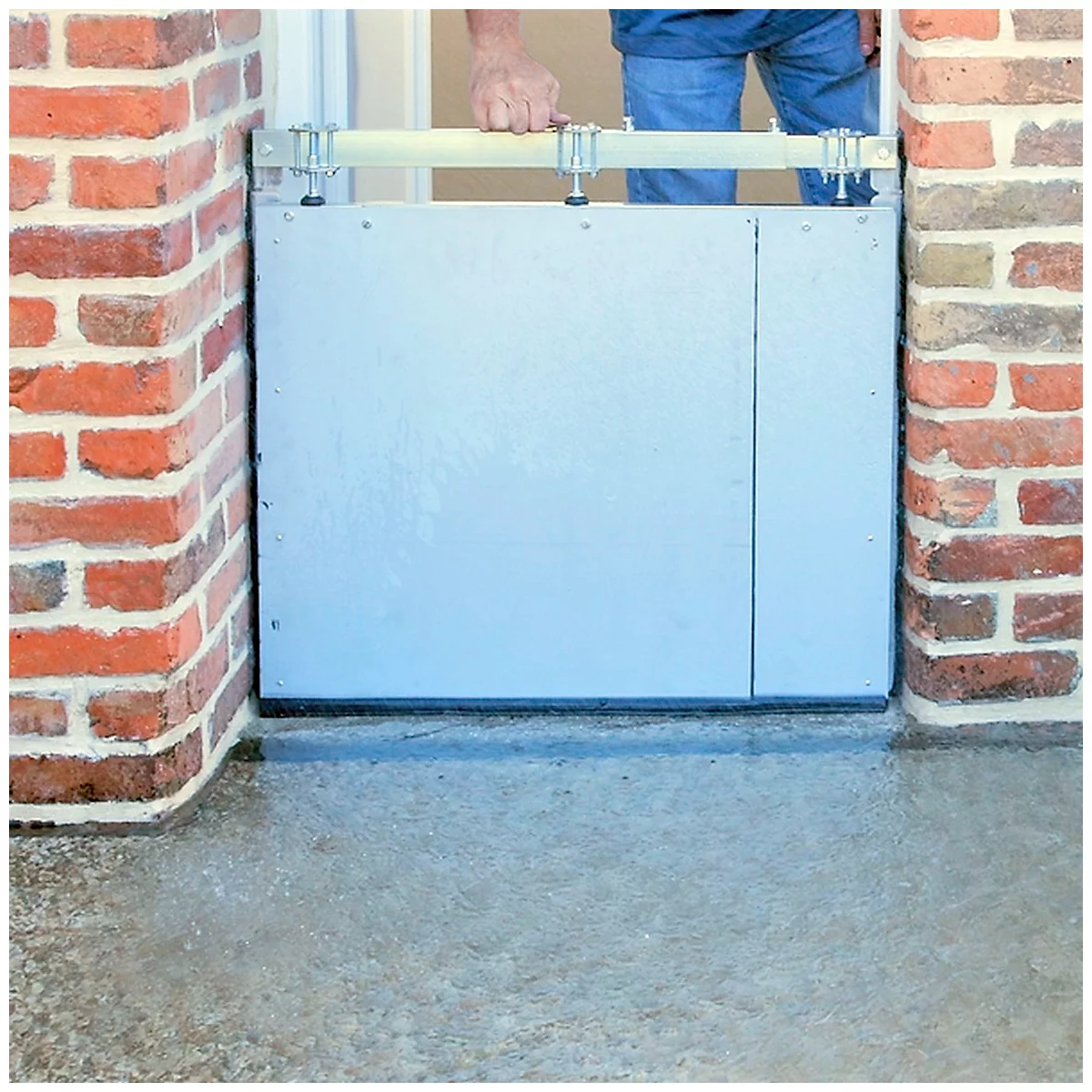 Une personne installe une barrière anti-inondation argentée devant une porte pour bloquer l'eau. Cadre en briques rouges.