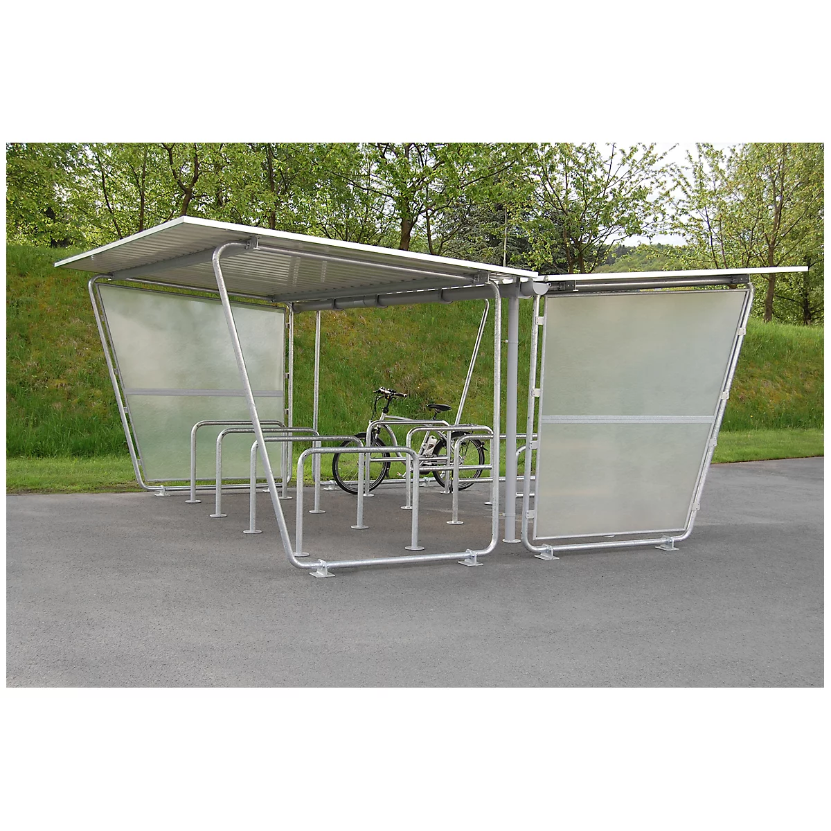 Bike shelter with bike. Metal canopy, transparent side walls. On asphalt, green trees in the background.