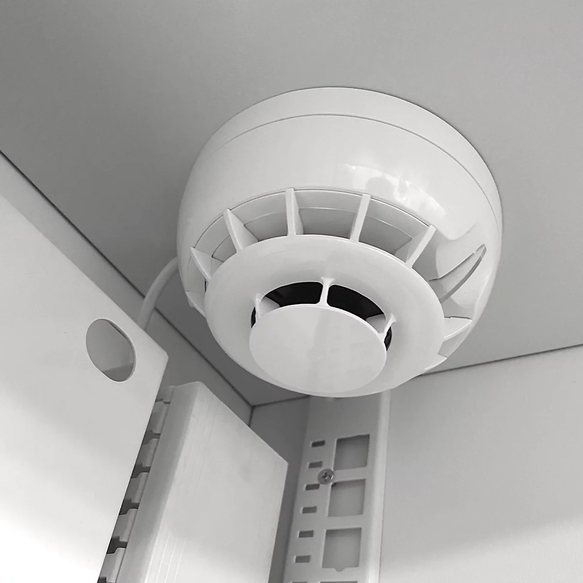 Weißer Rauchmelder an weißer Decke mit Lüftungsschlitzen. Kabel und Halterung sind ebenfalls zu sehen.